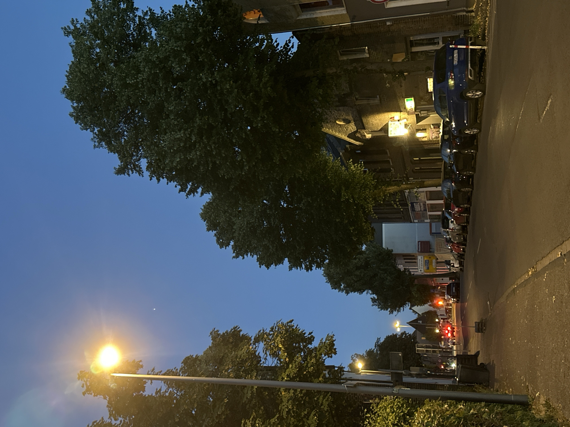 The image depicts a street scene in Krefeld, Germany, during twilight. A bright streetlamp illuminates the foreground, casting a warm glow on the sidewalk and the surrounding foliage. Several parked cars line the street, and a few vehicles are in motion further down the road, their brake lights visible. Buildings, appearing to be residential or commercial, flank the street, with some windows lit from within. The sky is a deep blue, transitioning from the day's light. Trees with lush green leaves are interspersed along the street, some casting shadows. The scene suggests a calm evening in a European city.