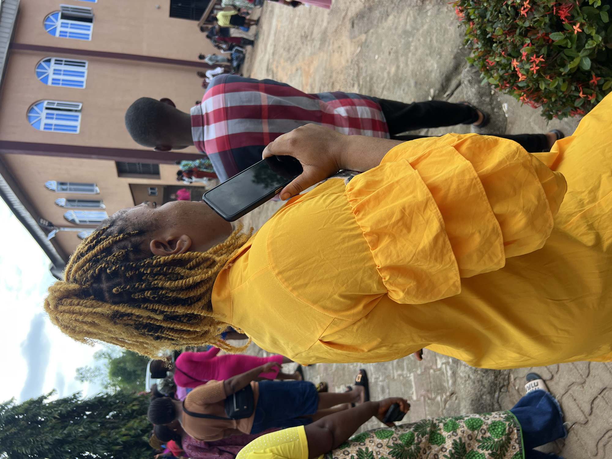 A medium shot shows a woman with blonde braided hair in a yellow shirt and holding a smartphone. She is standing outdoors with other people in the background, some of whom are wearing colorful clothing. The scene appears to be in front of a building with arched windows, possibly a church or community center, with a bit of greenery in the foreground. The location is identified as Nnewi, Nigeria.