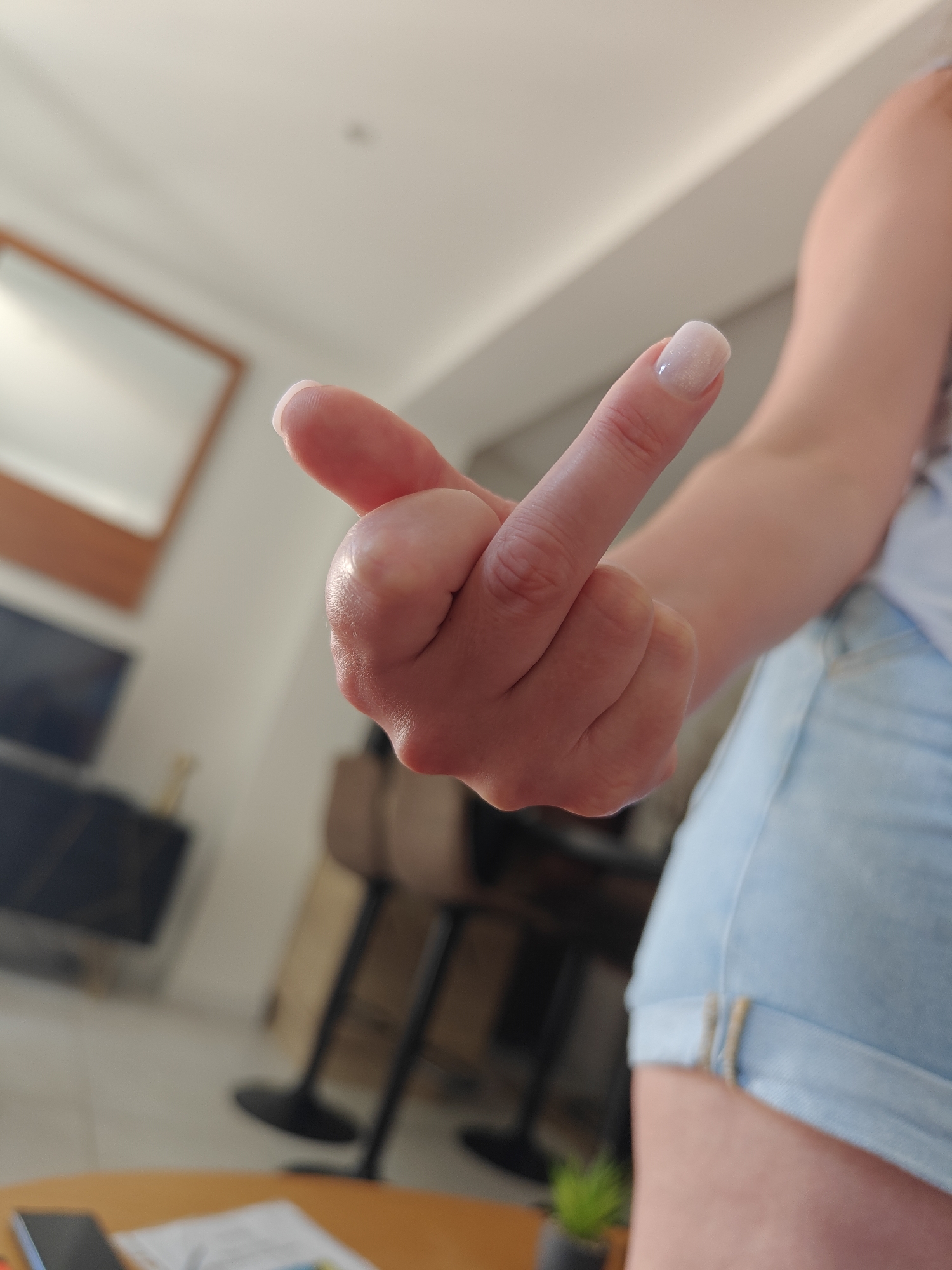 The image shows a close-up of a person's hand, with their middle finger extended upwards in a vulgar gesture. The hand has neatly manicured nails with a light, pearlescent polish. The person is wearing light-wash denim shorts. The background is softly blurred, suggesting an indoor setting. Visible in the background are the corner of a mirror with a wooden frame on a wall, a television screen mounted on a cabinet, and two bar stools. A wooden table is in the foreground, with a smartphone and some papers on it. The lighting suggests it is daytime. The overall tone of the image, with the gesture and the casual attire, conveys a sense of defiance or disrespect.