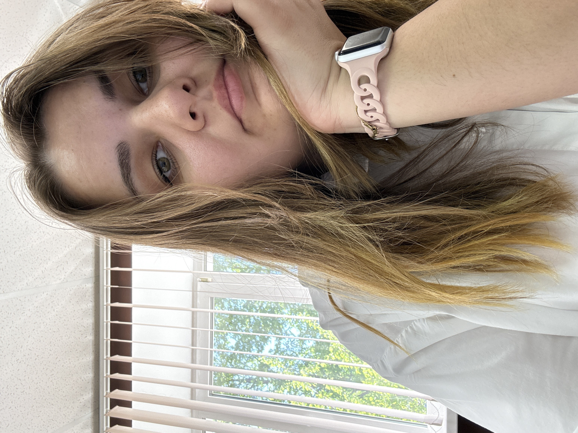 A selfie taken indoors, possibly in a room or office, with the subject being a young woman. She has light brown hair and fair skin, wearing a white shirt and a pink Apple Watch with a chain-link band. She is holding her hair with her hand. Behind her is a window with closed blinds, offering a view of trees outside, suggesting a residential or urban area. There is no information to suggest this photograph was taken in the city of Chaikovsky, Russia.
