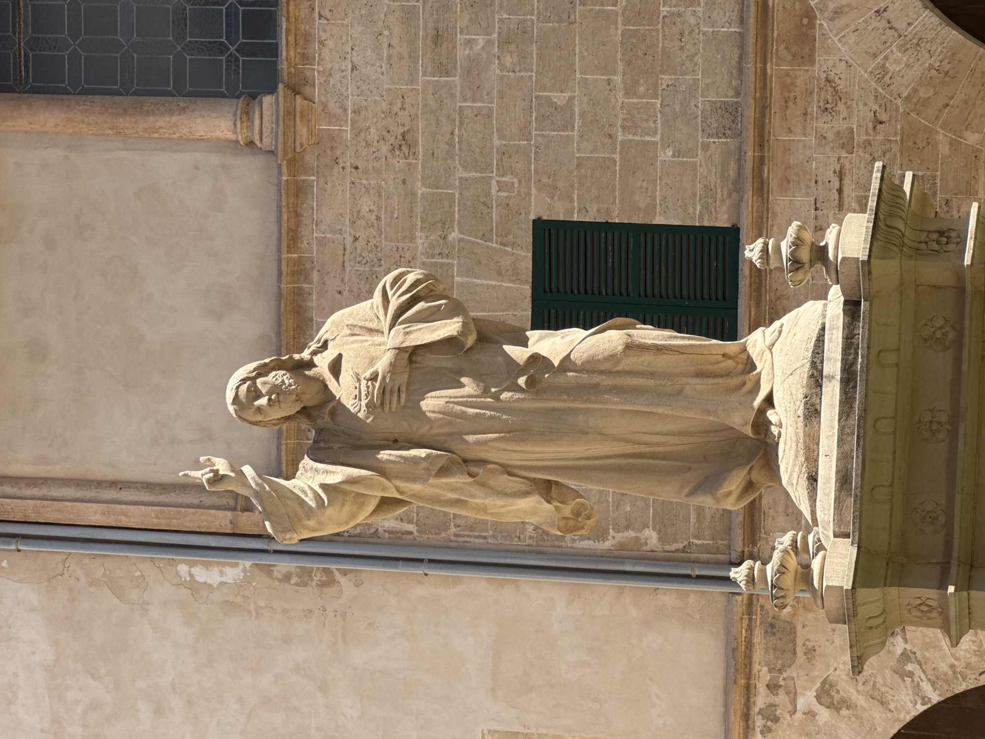 This is a medium-shot, eye-level, vertical photograph of a stone statue of Jesus Christ. The statue is made of light-colored stone and depicts Christ with a beard, long hair, and draped robes. He is standing on a pedestal, and his right arm is raised, with his index finger pointing upwards. His left hand is placed over his chest, near his heart. The statue is positioned in front of a building constructed of light brown brick. To the left of the statue, there is a light-colored textured wall with some staining, and two vertical grey pipes run down the wall. To the right of the statue, a dark green louvered window is visible against the brick wall, and above that, a darker, leaded glass window. The pedestal the statue stands on is ornate, with decorative carvings and urn-like finials on either side. The scene is bathed in bright sunlight, casting shadows. This statue is located in front of the facade of the Cathedral of Santa Maria of Palma, also known as La Seu, in Palma de Mallorca, Spain.