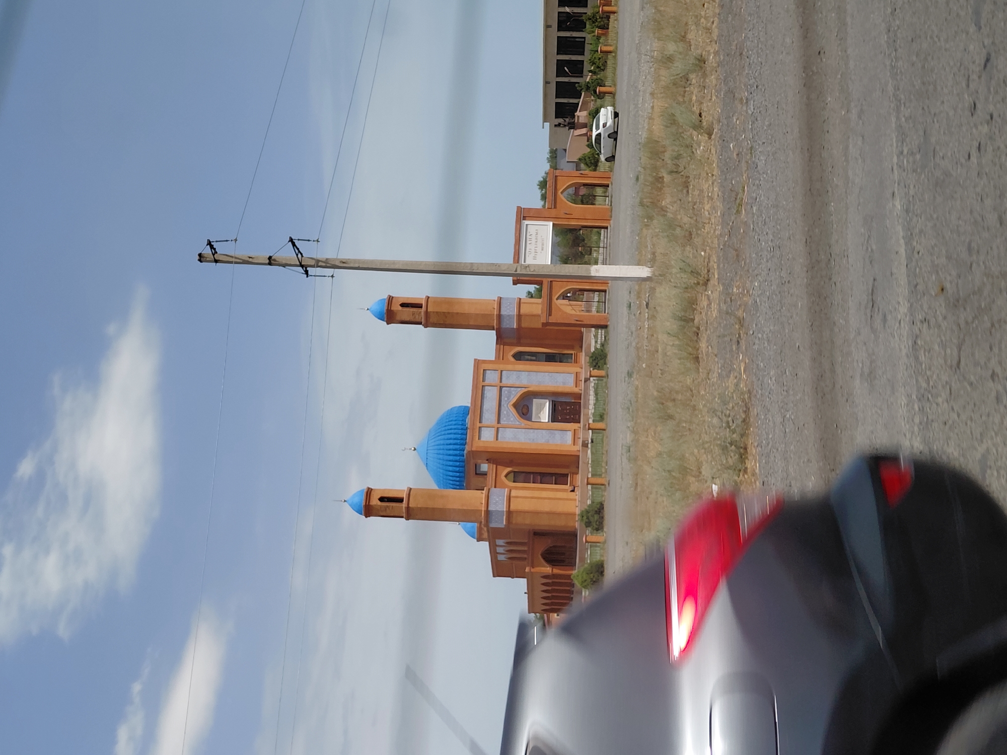 A mosque with a large blue dome and two smaller blue domes, flanked by minarets, is visible in the background under a partly cloudy sky. The mosque's facade is constructed of orange brick with blue tile accents. An archway entrance with a sign reading "ОЛ АНА" is positioned in front of the mosque. A white car is parked near the entrance. In the foreground, the rear of a dark-colored vehicle is blurred, suggesting it is in motion. The environment appears to be arid, with dry grass and gravelly terrain. The location is identified as Бұлақты, Kazakhstan.