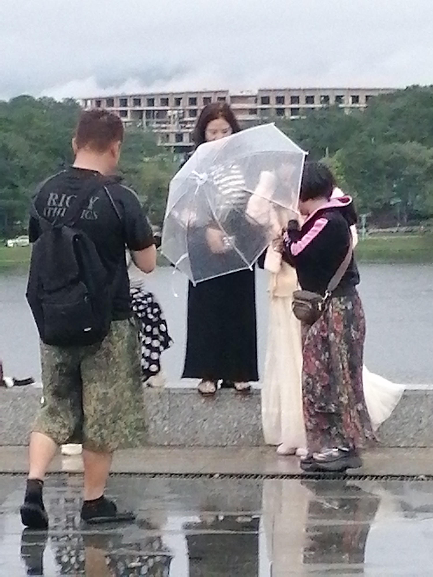 The image depicts a group of four people standing outdoors near a body of water. The scene appears to be slightly overcast, with a reflection of the surrounding environment visible on the wet ground.
In the foreground, a man is shown from the back, wearing a black t-shirt with "RICK Y ATHICS" printed on it, a black backpack, and camouflage shorts. He is wearing dark shoes.
To the man's right, three individuals are grouped together. One woman, wearing a long black dress, is partially obscured by a clear umbrella. Another woman is wearing a long, light-colored dress and standing next to her. The third person, also a woman, is wearing a black jacket with pink trim over a floral-patterned long skirt and has a small brown bag slung across her body. She is also wearing dark, platform shoes.
In the background, there are trees and a large, multi-story building under construction. The overall setting suggests an urban or semi-urban area, possibly a park or waterfront promenade.
Based on the presence of extensive construction and the architectural style visible in the background, this scene could potentially be located in Da Lat, Vietnam, a city known for its ongoing development projects and natural beauty.