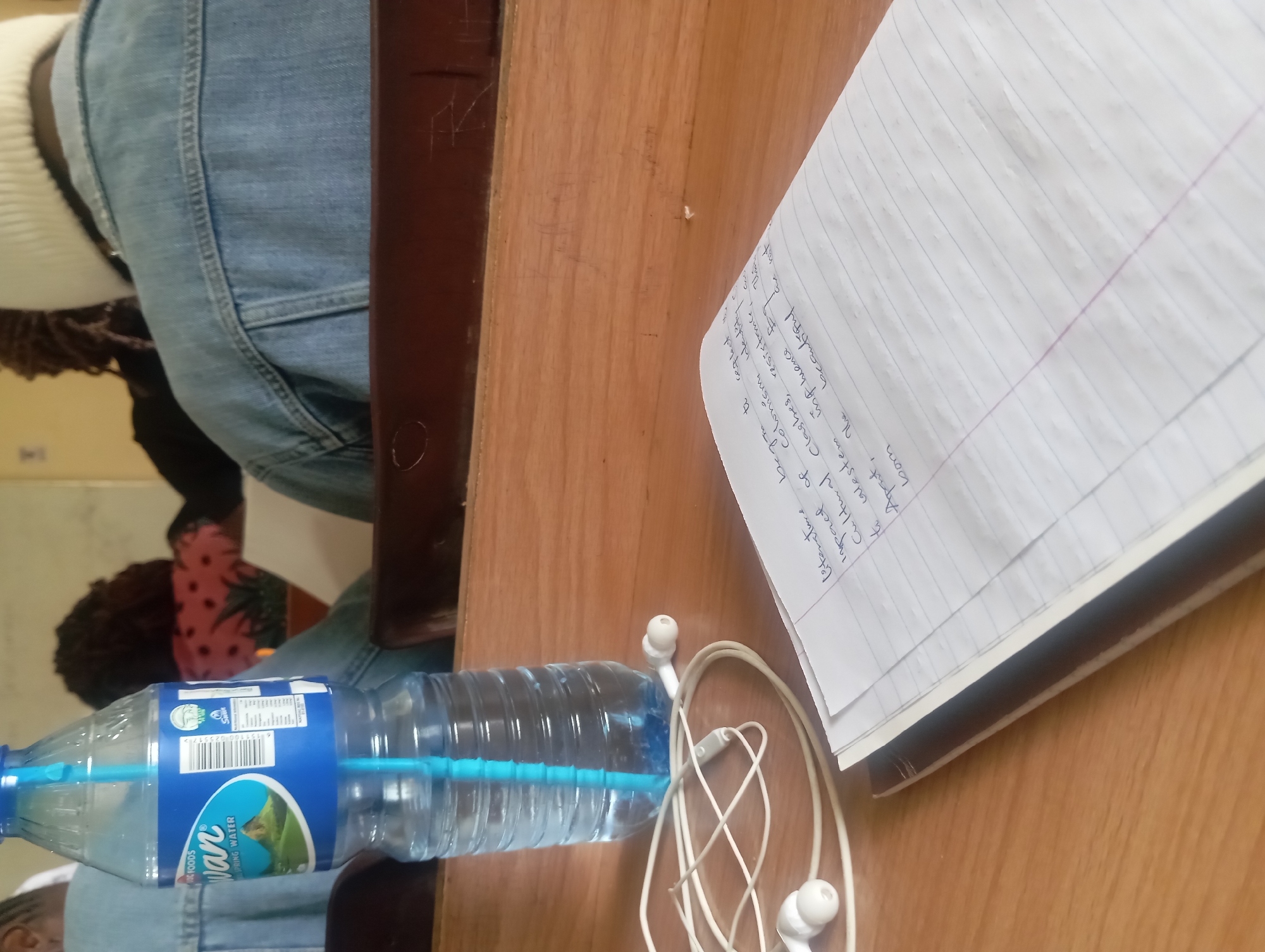 A high-angle, medium shot captures a desk in what appears to be a classroom. In the foreground, a clear plastic bottle of "Swan" brand spring water stands upright, its blue straw visible inside. To the left of the water bottle, a pair of white earbuds with their cord are loosely arranged on the wooden surface.
In the center of the frame, a notebook with lined paper is open, displaying handwritten text in dark ink. The writing discusses "literature," "impact of colonialism," "cultural clashes," "resistance," and "western influence." Some of the text reads: "Literature began to reflect the impact of colonialism, identity, cultural clashes, resistance... western influence. The 'aperture,' the beautiful bloom."
In the background, out of focus, are the backs of several people. One individual on the left wears a blue denim jacket. Another person in the center wears a blue denim jacket and a white beanie. To the right, partially visible, is another person in a denim jacket. The environment suggests a public space, likely an educational setting, given the presence of students and writing materials.