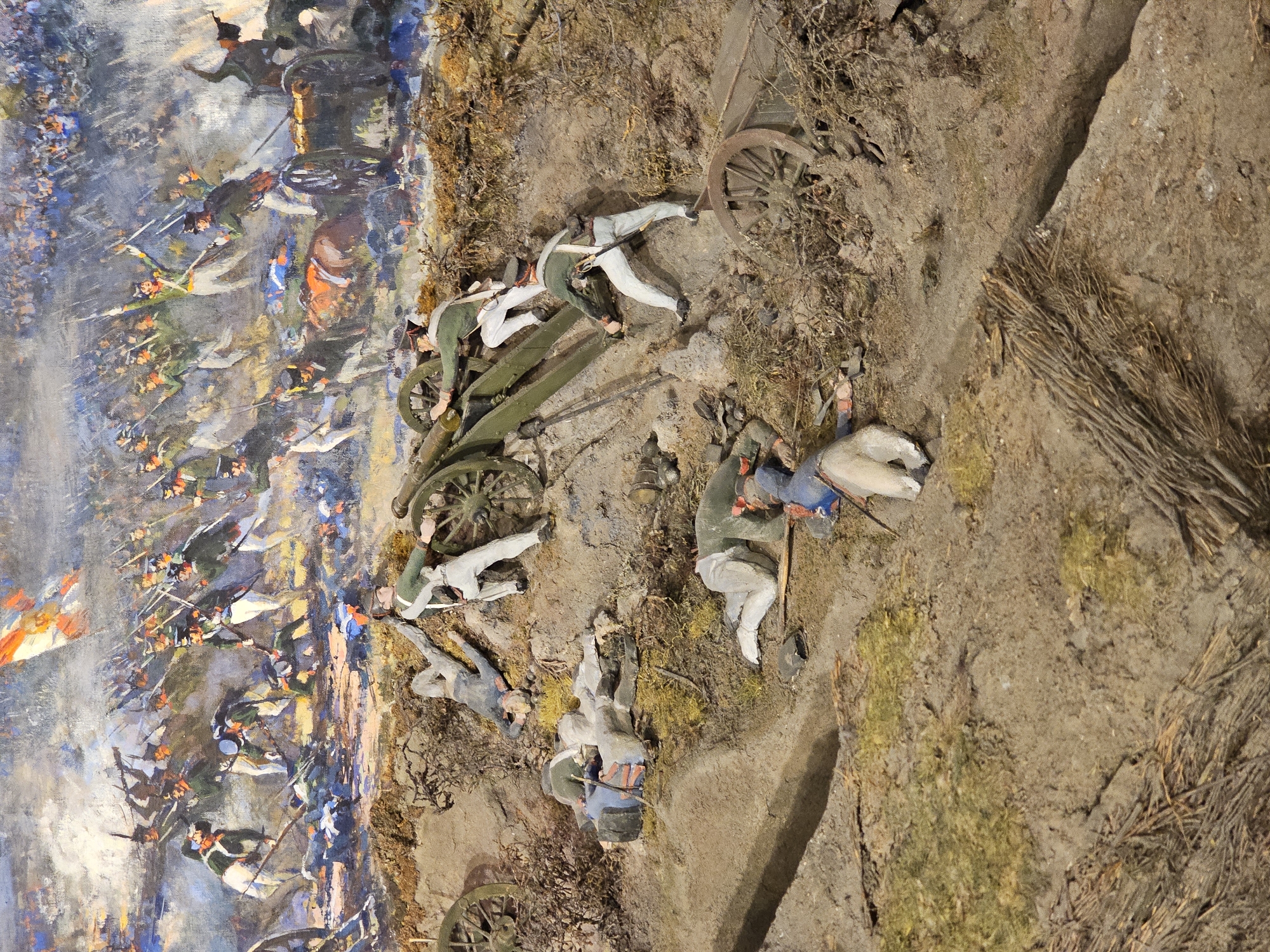 The image depicts a diorama representing a battle scene, potentially the Battle of Borodino, fought near the city of Borodino, Russia. The diorama features miniature figures of soldiers, some fallen, around a cannon. A painted backdrop shows a larger battle scene with more soldiers and flags. The ground is modeled with earth, twigs, and vegetation.