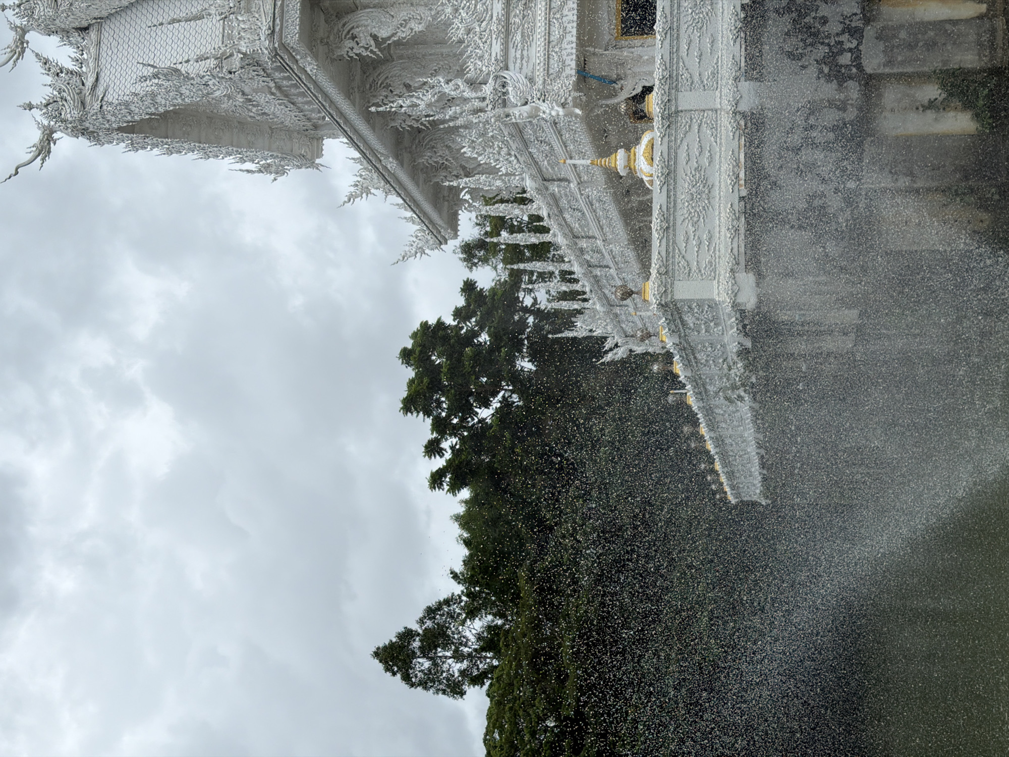 The image shows the White Temple (Wat Rong Khun) in Thep Krasatti, Thailand, partially obscured by a heavy downpour. The upper section of the ornate white temple structure is visible against a cloudy sky, with intricate carvings and gold accents. Lush green trees are positioned below the temple roofline. A dense curtain of rain fills the lower portion of the image, obscuring the surrounding environment. The water appears to be falling into a body of water below.