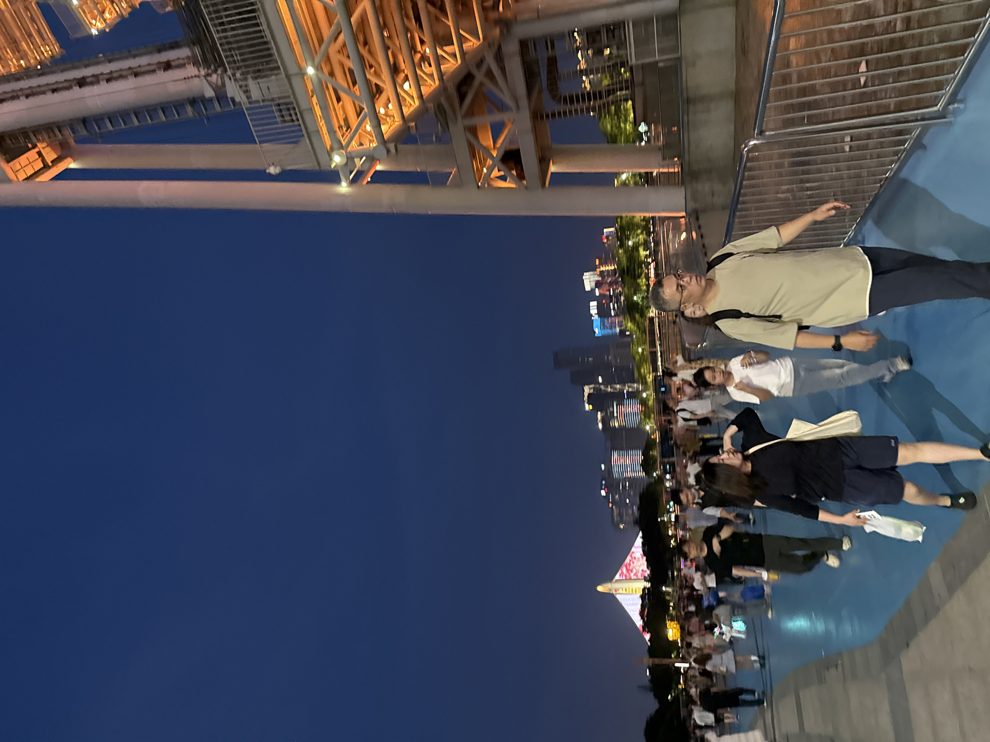 The image shows a group of people walking along a waterfront promenade in the Tianhe District, China, at dusk. The promenade has a blue surface, and a metal railing separates it from a lower level. In the foreground, a man with glasses and a backpack walks towards the right. To his left, a woman in a black shirt and dark shorts carries a white bag. Further behind them, other individuals and groups are visible, walking in the same direction or in different directions. In the background, a modern city skyline is illuminated by lights, with a distinctive building featuring a white spire and a colorful, patterned facade. To the far right, a large, illuminated structure with a complex metal framework, possibly part of a bridge or tower, dominates the frame. The sky is a deep blue, indicating the transition from day to night.