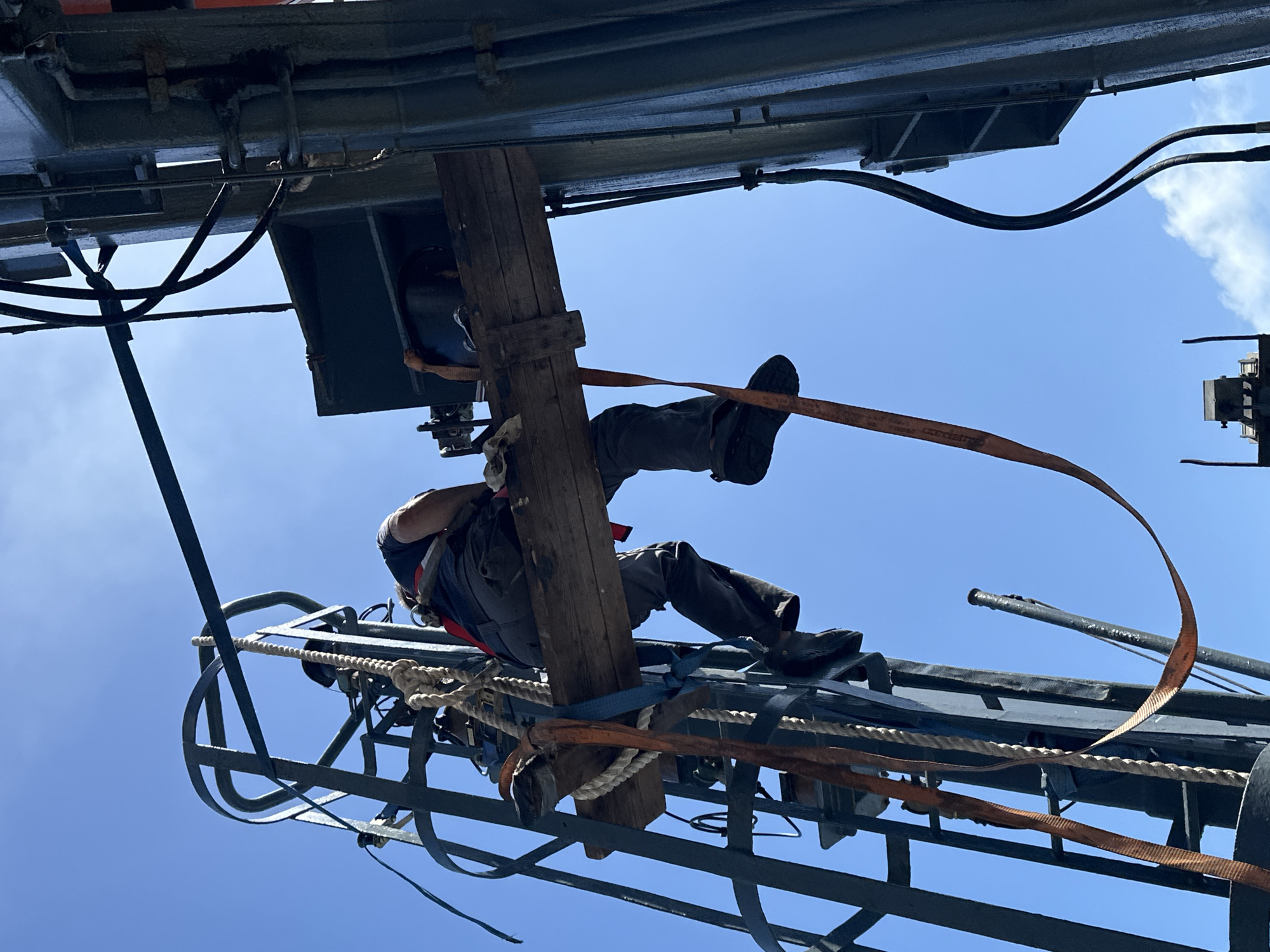 The image depicts a worker suspended in the air, secured to a complex structure. The scene is oriented upward, with a clear blue sky in the background. The worker is wearing a harness and appears to be attached to both a wooden beam and the metal structure. There are numerous ropes, cables, and straps visible, suggesting a high level of complexity in the rigging. The metal structure features a ladder-like section. The location in the city of Unknown, Unknown is unknown.