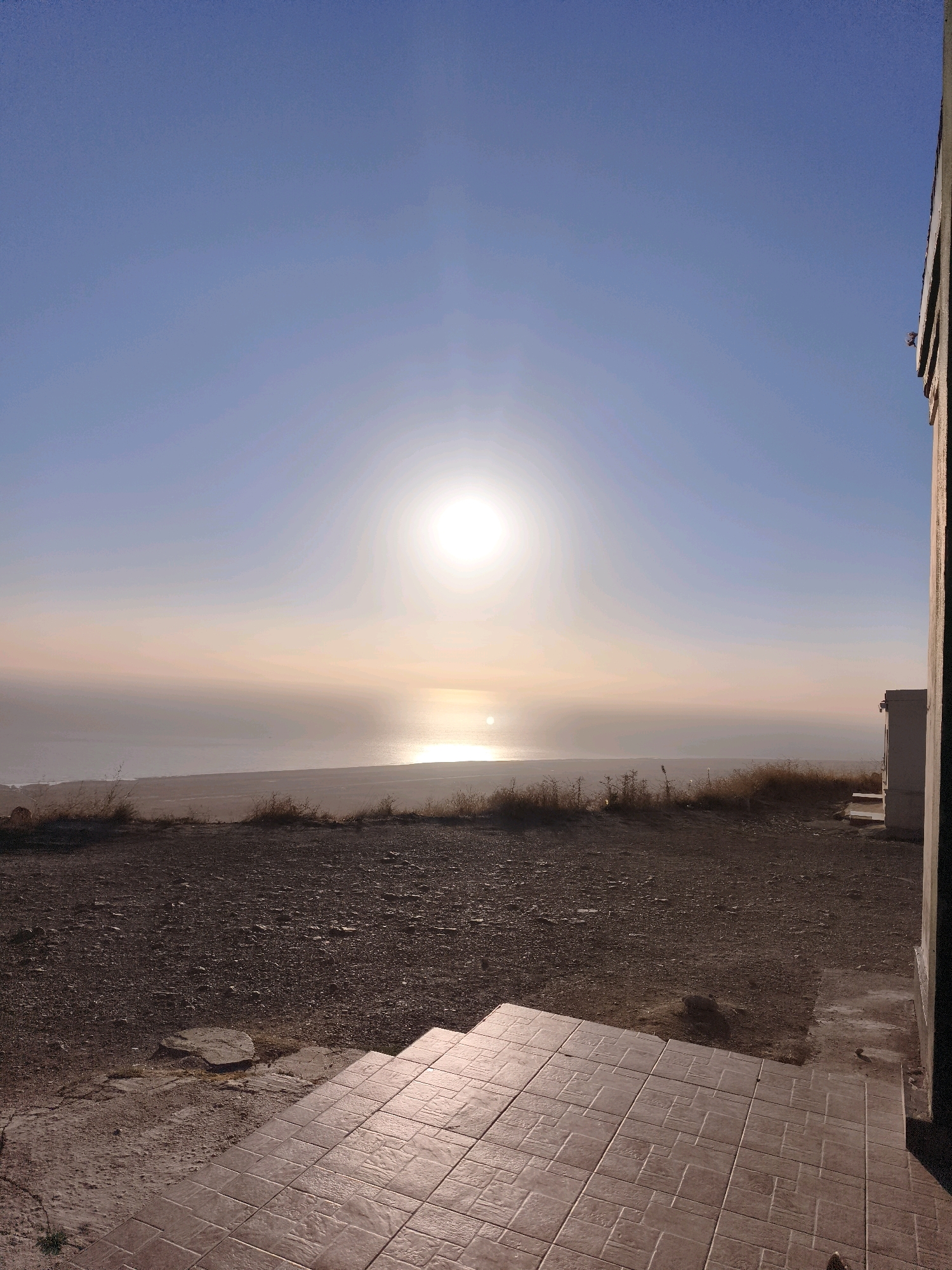 The image depicts a natural landscape at what appears to be sunrise or sunset. The sun is prominently visible in the sky, casting a bright, hazy light across the scene. A body of water, likely the sea or a large lake, is visible in the distance, with a reflective glint on its surface. The foreground is dominated by a rough, uneven terrain covered in gravel and dry vegetation, suggesting an arid or semi-arid environment. To the right of the image, a portion of a building structure is visible, with a light-colored wall and what appears to be a doorway or opening. The sky is a clear, pale blue, transitioning to softer hues near the horizon. There are no people or animals visible in the image. The scene is likely located in a coastal or lakeside area, possibly a relatively undeveloped or rural part of Qalaşıxı, Azerbaijan, given the arid terrain and natural elements.