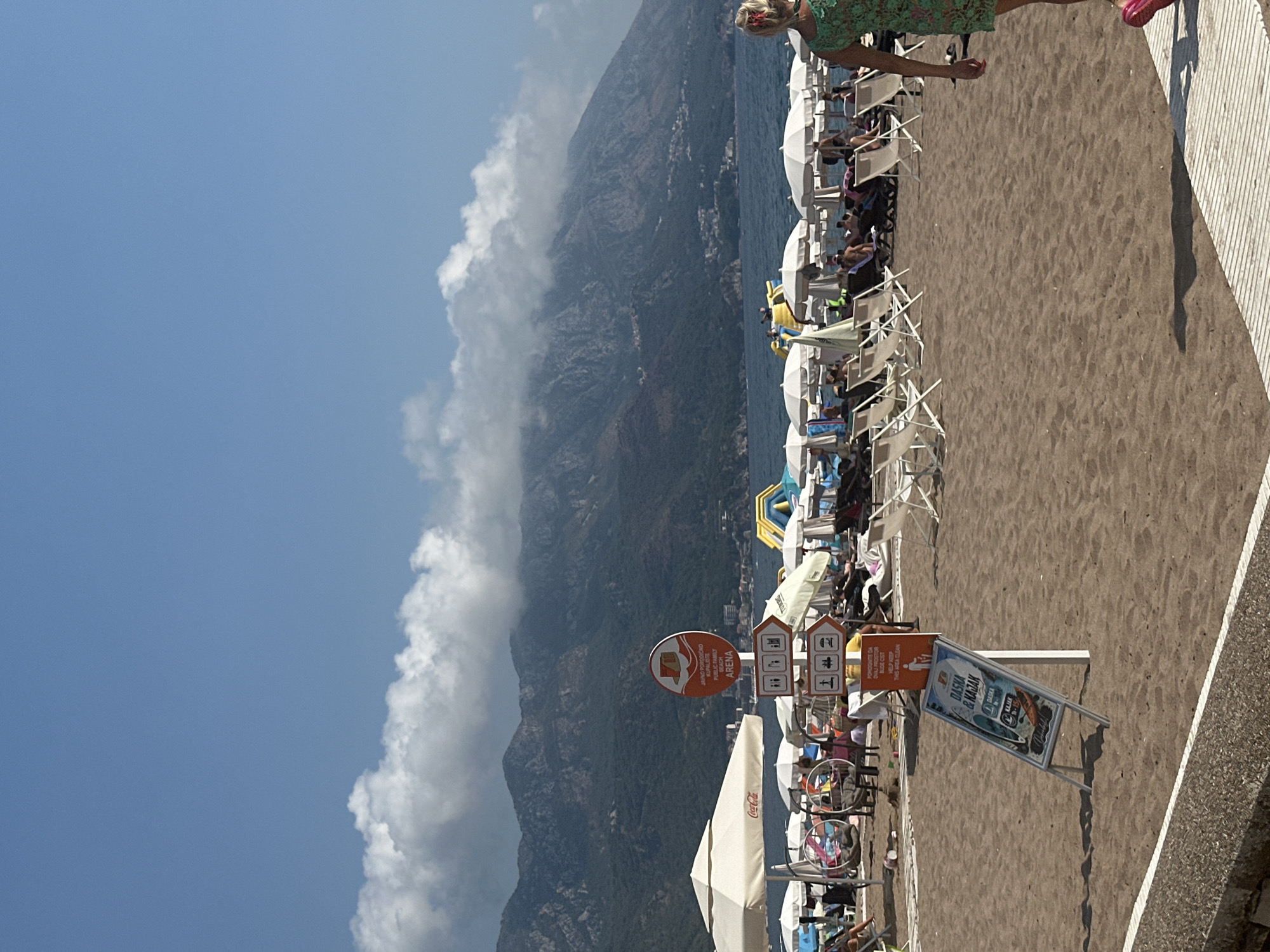 The image shows a crowded beach in Boreti, Montenegro, on a sunny day. The sand is dark brown, and numerous white beach umbrellas and lounge chairs are set up. In the background, the dark blue sea meets a chain of lush green mountains partially obscured by a thick band of white clouds. Several people are visible, lounging on chairs or walking on the beach. One woman, wearing a green dress and pink shoes, is walking away from the camera towards the right side of the frame. Signs indicating directions to "ARENA" and various beach activities like "Daska & Kajak" (board and kayak) are present. A yellow and blue inflatable water slide is visible in the sea. The overall scene depicts a typical summer day at a popular beach resort.