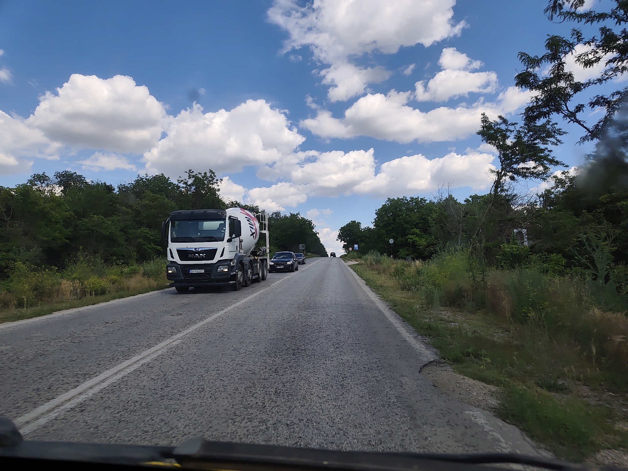 A white cement truck, identified as a MAN vehicle by its emblem, is traveling on a paved road with a double white line marking the center. The truck has a partially visible Bulgarian license plate that reads "EH 9165 KT". Behind the cement truck, two black cars are also on the road, followed by another vehicle further in the distance. The road is lined with lush green trees and vegetation on both sides. The sky is a vibrant blue, dotted with fluffy white clouds. This scene is consistent with a location on the outskirts of Pleven, Bulgaria, likely on a route leading out of the city, given the mix of road infrastructure and natural surroundings. The perspective suggests the image was taken from inside another vehicle, looking forward.