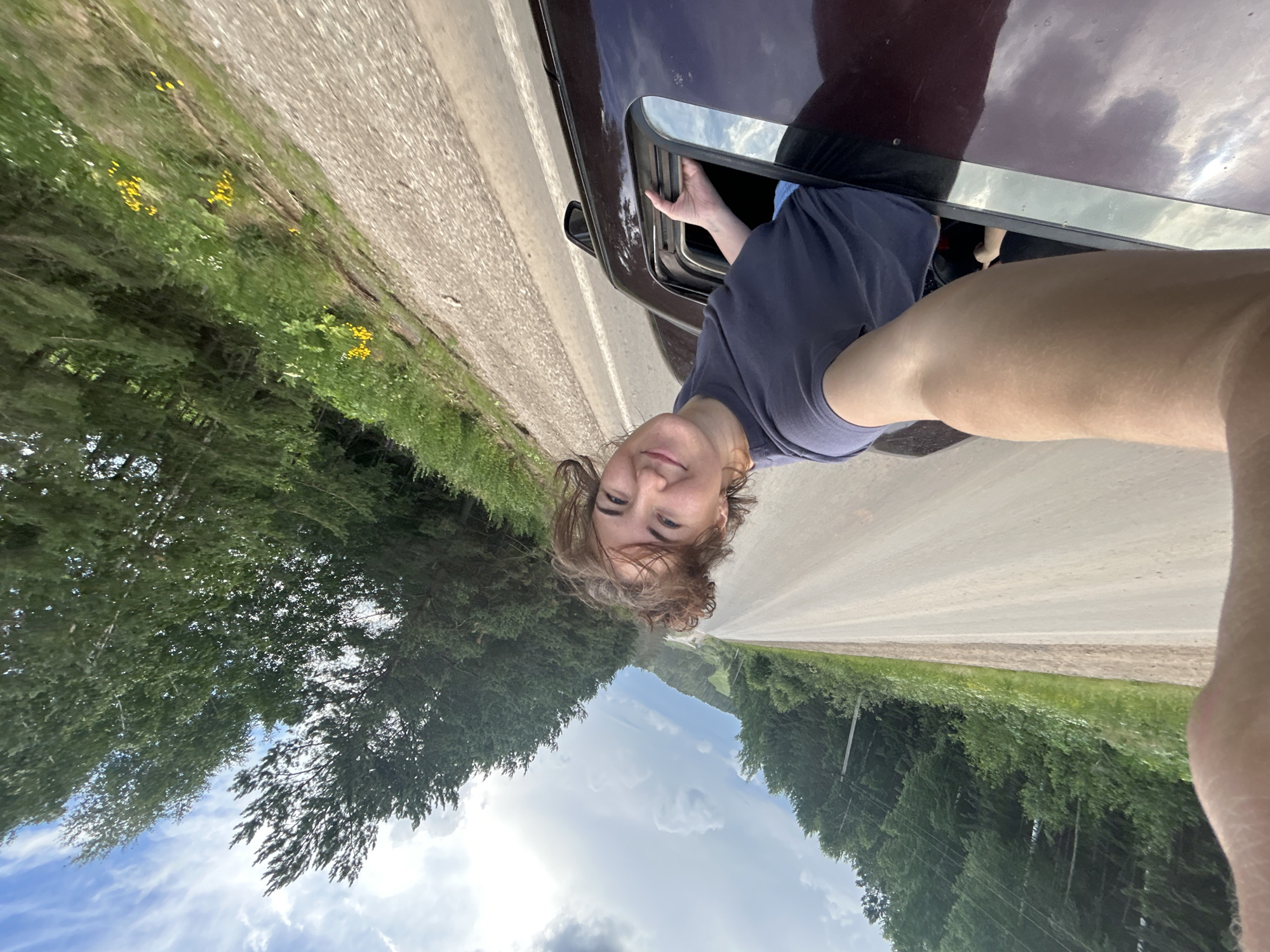 A young woman with messy brown hair, wearing a dark blue t-shirt and denim shorts, leans out of a car sunroof while smiling and holding the frame. Her left arm is extended, holding the camera for a selfie. The car appears to be on a paved road with a gravel shoulder, bordered by a dense forest of green trees and some yellow wildflowers. The sky is partially cloudy with patches of blue. The image is taken from a high angle, looking down and to the right. The context of the city of Верх-Рождество, Russia is inferred from the provided information, though not explicitly visible in the image itself.