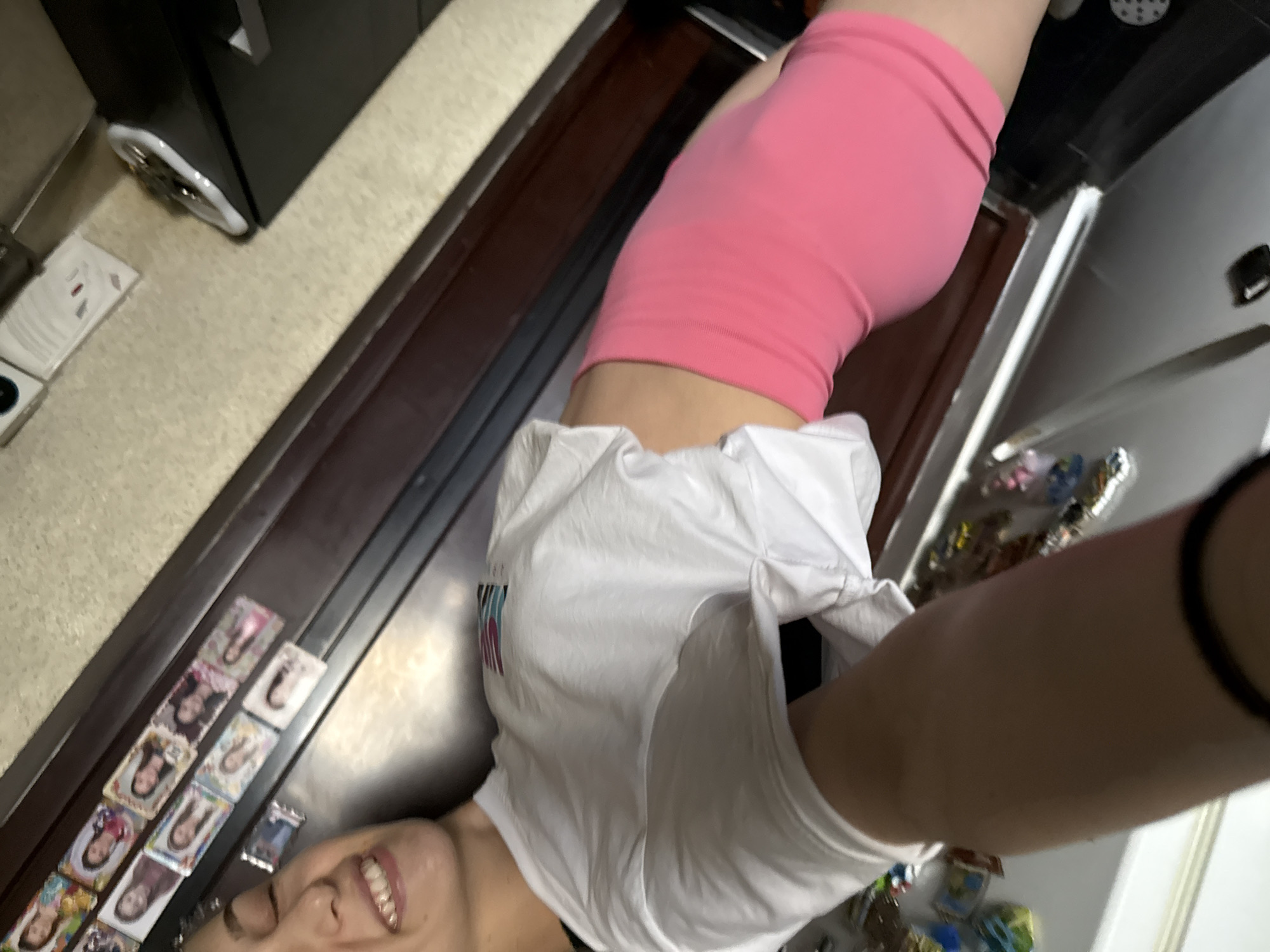 A selfie depicts a person wearing pink shorts and a white t-shirt, taken at an unusual angle. A counter or table is covered with collectible cards or stickers, and the background includes a refrigerator adorned with magnets. A kitchen appliance is visible on the left. It is impossible to determine if the image was taken in Sofia, Bulgaria from the visual data provided.
