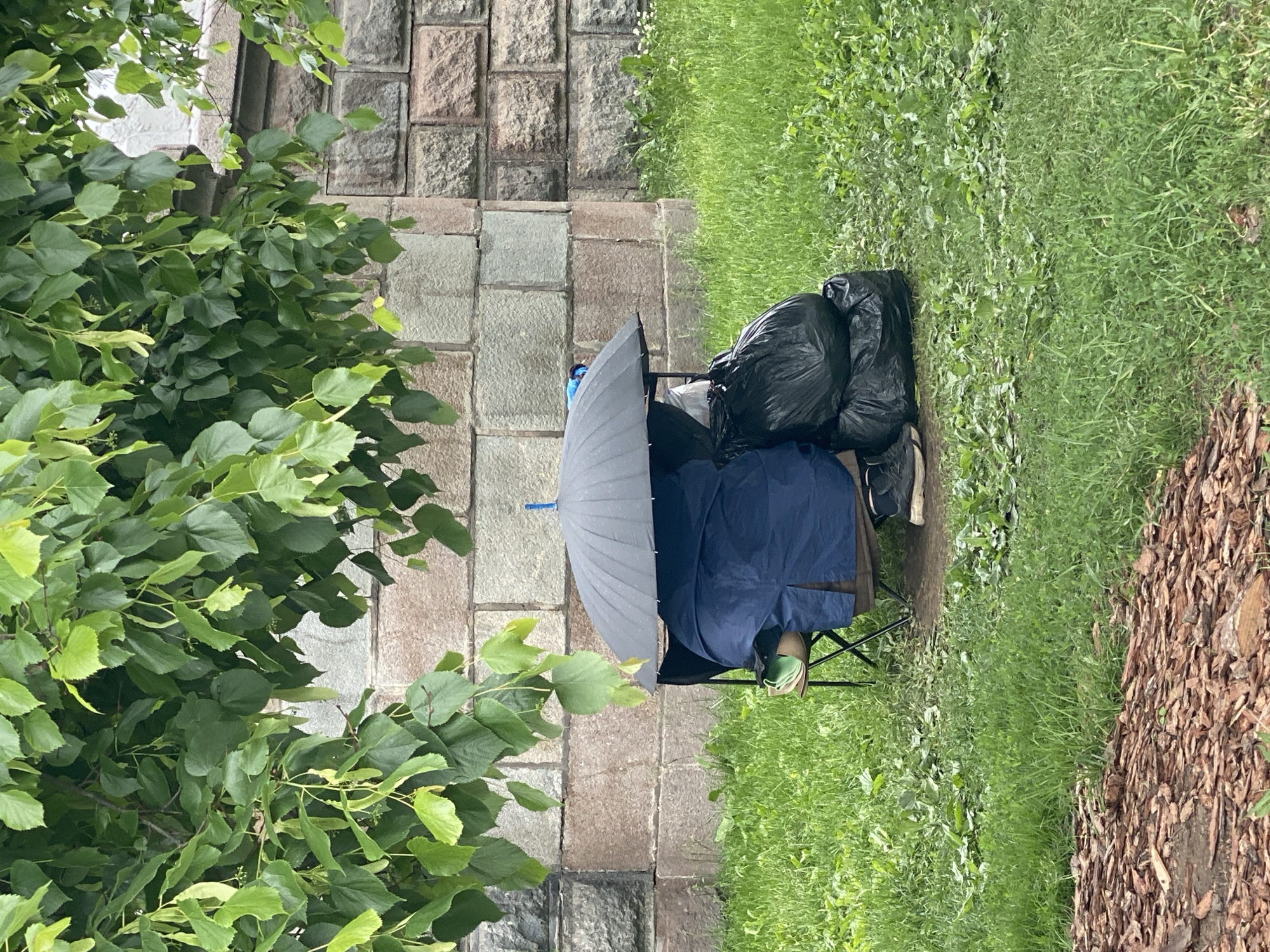 A scene in Moscow, Russia depicts a setup against a stone wall and adjacent to grass and mulch. A person sits in a black folding chair under a large grey umbrella with a blue handle, also covered by a navy blue tarp or garment. Behind the chair are two large black garbage bags, along with a pair of grey sneakers. The ground cover transitions from green grass to mulch or wood chips to the right. A tree with green leaves is positioned on the left side of the frame. The stone wall behind is constructed of large, rough-cut blocks.
