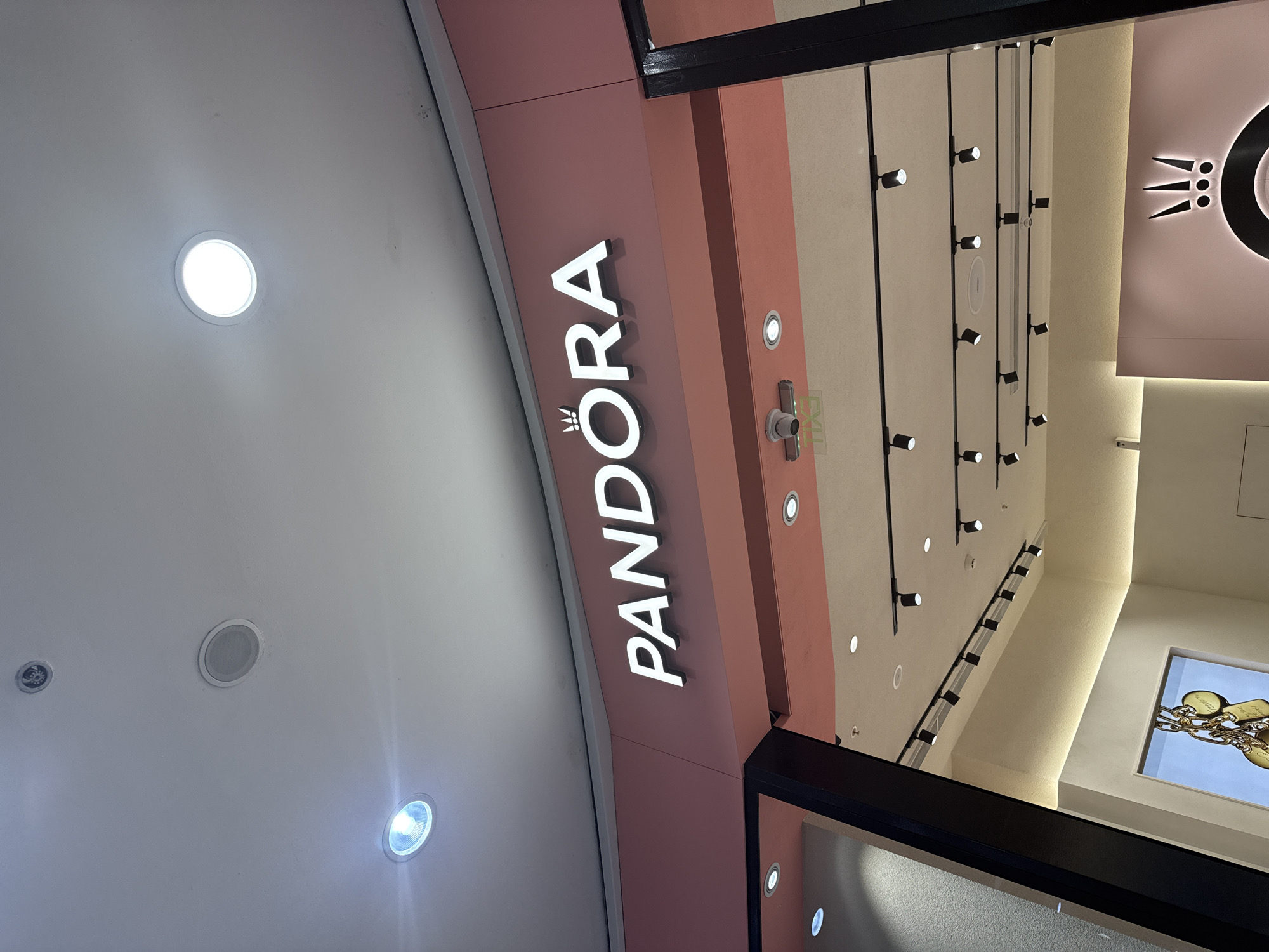 The image shows the entrance to a Pandora store. The store's sign is vertically oriented on a pink wall, displaying the "PANDORA" logo in white letters. Above the entrance, there are several recessed lights on the ceiling. The interior of the store features a variety of lighting fixtures and a display of jewelry. The location is within an urban area, but the city and precise location are unknown.