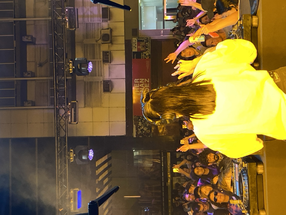 A nighttime scene depicts an outdoor DJ performance in Buenos Aires, Argentina. The foreground is dominated by a person, seen from the back, with long dark hair, wearing a bright yellow long-sleeved shirt and black Technics over-ear headphones. This individual is positioned behind dark DJ turntables and a mixer, actively engaged in a performance. Behind the DJ, a dense crowd of people fills the mid-ground. Numerous individuals in the audience hold up smartphones, recording or photographing the event, while others have their hands raised in the air.
The background consists of a multi-story urban building facade, featuring a grid of windows and several external air conditioning units. Directly behind the audience, a red banner with white text reading "HERNAN DEZ" is visible, along with the street number "2138," indicating a specific address likely on Pellegrini street in Buenos Aires. Above the performance area, a metal truss supports multiple stage lights, some emitting blue and purple illumination into a visible atmospheric haze. The setting is an urban street or open-air venue, characterized by artificial lighting and an energetic, engaged crowd.