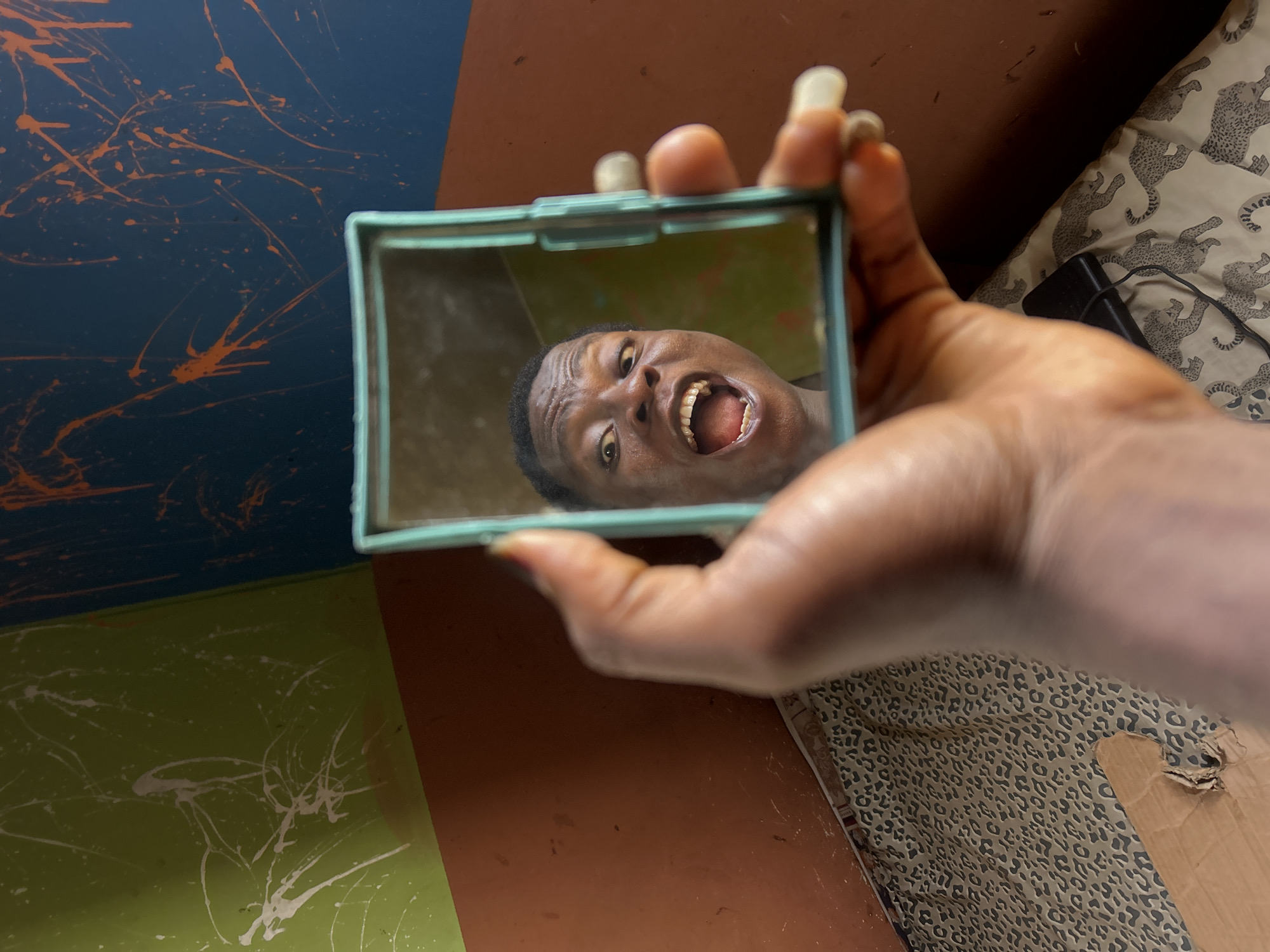 A close-up, high-angle shot captures a dark-skinned hand holding a small, rectangular mirror with a light blue frame. The hand has long, light-colored fingernails. Reflected prominently in the mirror is the face of a person, likely the individual holding the mirror, with dark skin and short, dark hair. The person's face displays an exaggerated expression, with wide-open eyes and a widely opened mouth revealing teeth and tongue, suggesting surprise, shouting, or a playful grimace.
The background consists of a multi-colored wall or surface. To the upper left, there's a dark blue section with orange paint splatters. Below it is a green section with white paint splatters. A solid brown-orange section runs along the right side and bottom. In the lower right corner, behind the hand, a leopard-print fabric, possibly bedding or a blanket, is visible, along with a black rectangular object resembling a power adapter or charger. The scene suggests an indoor setting, possibly a bedroom, and depicts a moment of expressive self-reflection or playful interaction with the mirror. No visible text is present.