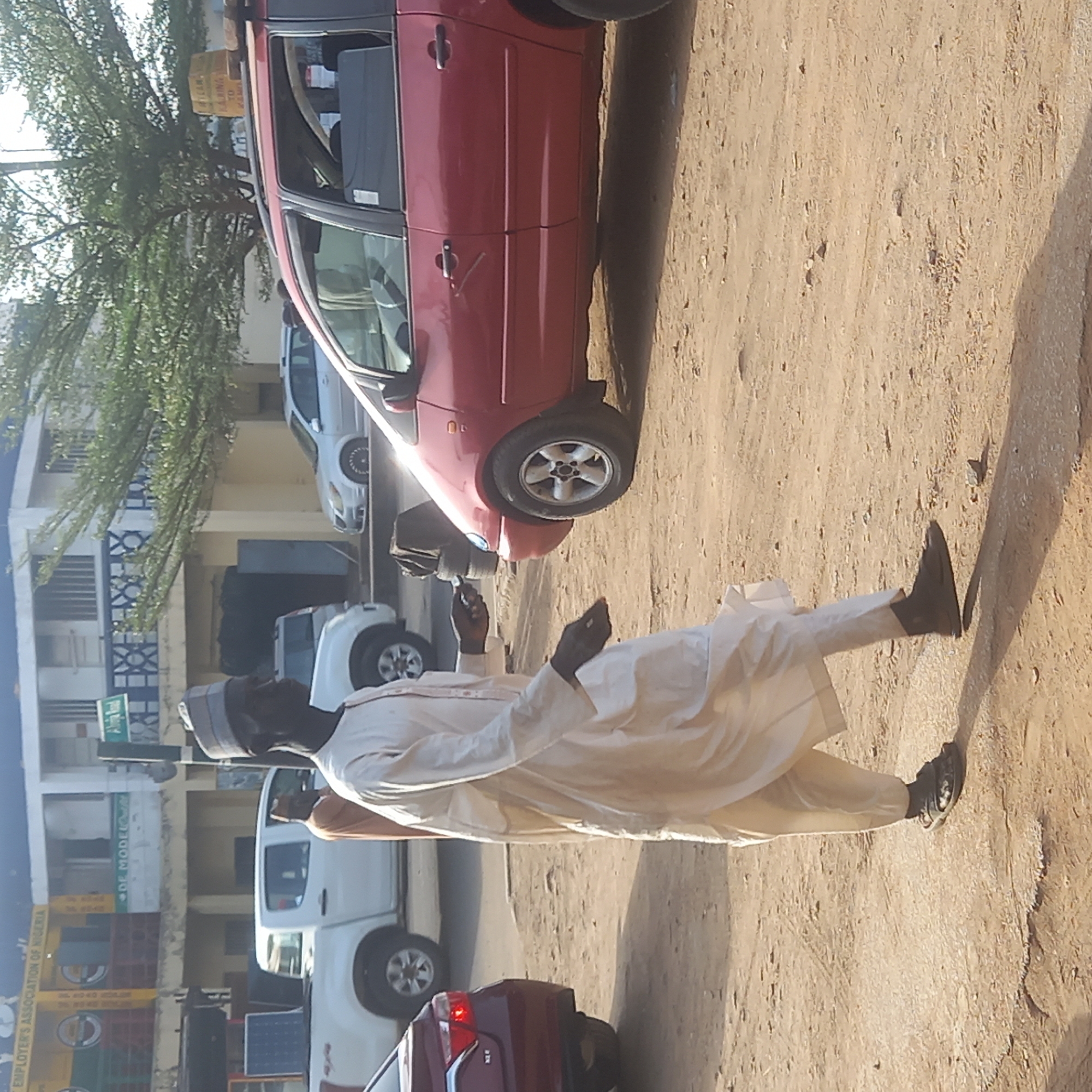 This image captures a daytime street scene in what appears to be an urban or semi-urban area, likely Dadi Riba, Nigeria, given the contextual information. The photograph is taken from a slightly low angle, looking across a dirt or unpaved road.
The primary subject is a man in the foreground, walking towards the left of the frame. He is dressed in traditional light-colored attire, possibly a jalabiya or agbada, and wears a matching cap. He has dark-colored shoes and appears to be holding a small dark object, possibly a phone, in his left hand. His right arm is slightly extended. Long shadows are cast on the ground to his right, indicating strong sunlight, likely in the late afternoon or early morning.
Behind the walking man, a red sedan or hatchback is prominently parked on the unpaved road. Its wheels and side profile are clearly visible. Further in the background, a white pickup truck is parked, and another white sedan is partially visible behind the red car. Buildings line the left side of the street, featuring architectural details such as balconies and patterned railings. Green trees with abundant foliage are also visible behind the buildings.
Visible text includes a green street sign that partially reads "Dadi Road". On a yellow banner or sign attached to one of the buildings, text like "THE CITY'S ASSOCIATION OF NIGERIA" and "DF MODEL" can be deciphered, along with what appear to be phone numbers starting with "080". Stacked yellow and blue crates are also seen near one of the buildings, and a small solar panel lies on the ground near the white pickup truck. The overall impression is a bustling street scene under clear, sunny weather.