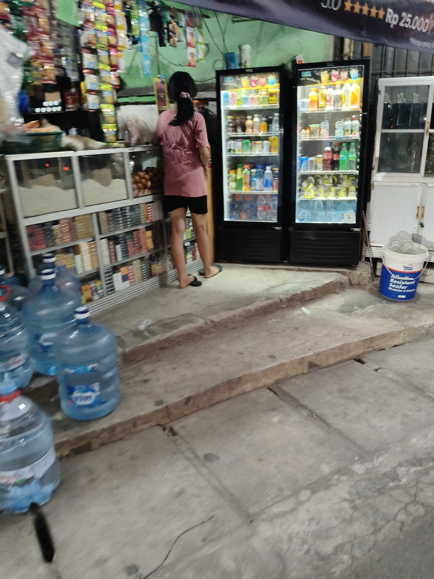 The image depicts an outdoor convenience store setting, likely in South Tangerang, Indonesia, given the context of such establishments in the region. In the foreground, several large, clear plastic jugs of drinking water are arranged on a concrete platform. To the left, a person wearing a pink t-shirt and black shorts stands facing away from the camera, browsing items displayed on shelves behind a glass counter. Behind the person, shelves are filled with various packaged goods. In the background, two illuminated glass-fronted refrigerators are stocked with beverages. To the right of the refrigerators, a white cabinet with a glass door stands open, and a blue and white bucket containing several clear glass cups is placed on the ground. The scene appears to be from an evening or nighttime setting, as indicated by the lighting from the refrigerators and the overall ambiance. The concrete ground shows signs of wear and tear.