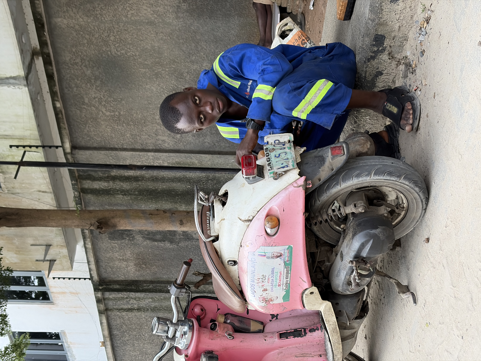 A young Black male wearing a blue and yellow jumpsuit is sitting on a curb next to a pink and white scooter in Igboefon, Nigeria. The man is looking at the camera. The scooter has a sticker on its side with the text "LAGOS MAINLAND" and a picture of a man. The man is wearing black sandals. The background shows a grey wall, a tree, and a building.