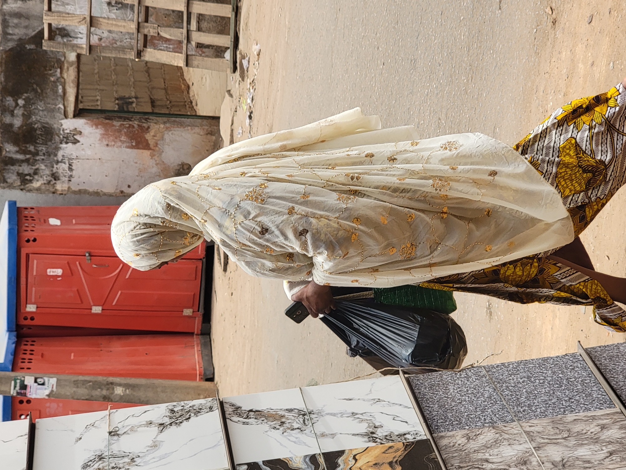An individual in Maiburiji, Nigeria, dressed in a white head covering and a patterned dress, is seen holding a black plastic bag and a mobile phone. They are standing next to a display of tiles outside a building where orange portable restrooms are stacked. The background includes a building facade with exposed wooden beams and a dirt or paved road.