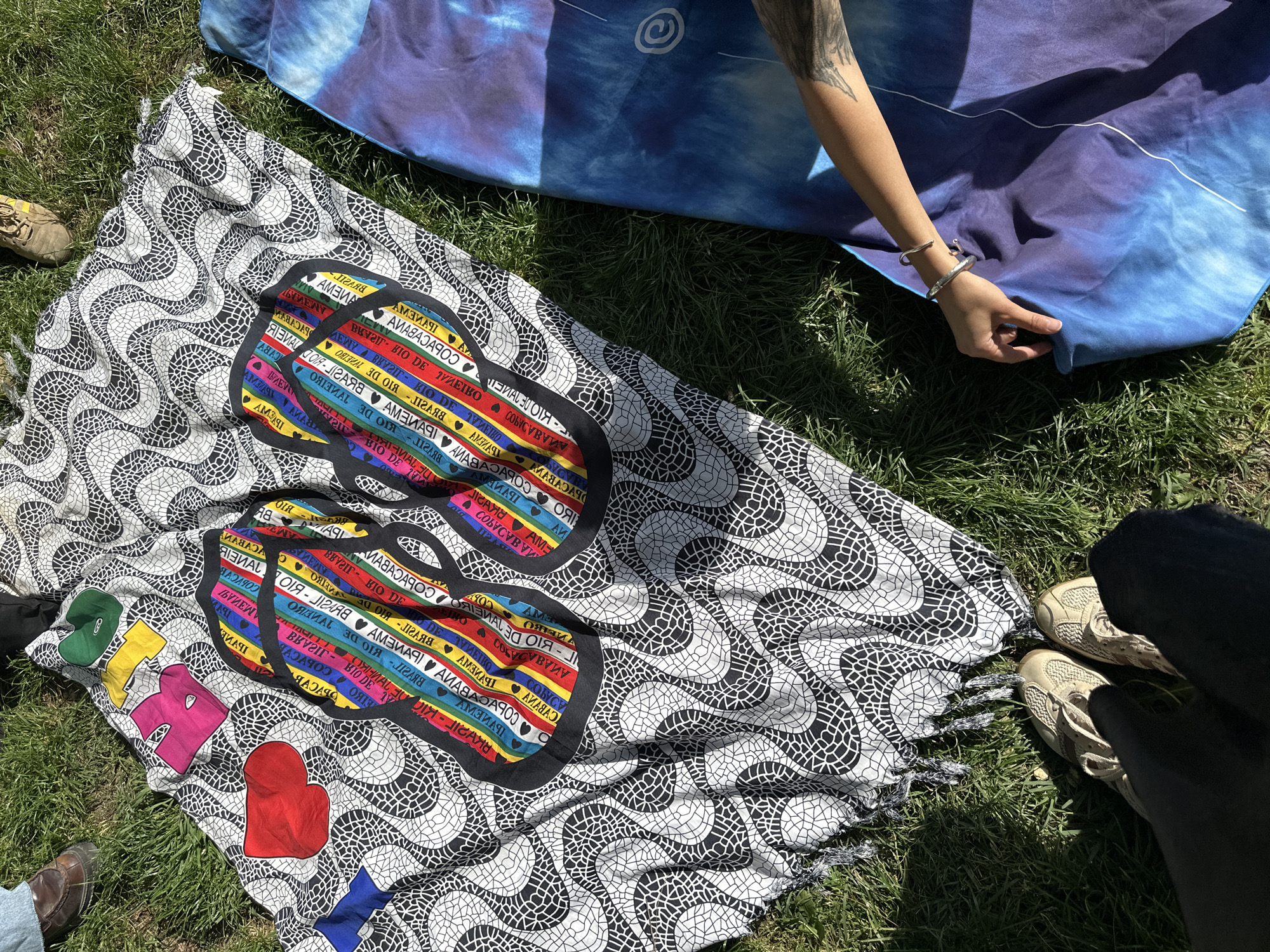 The image shows a grassy area with two patterned blankets spread on the ground. The top blanket is predominantly blue with a spiral design. A person’s arm, adorned with a tattoo and bracelets, is visible, holding the edge of the blue blanket.
Below the blue blanket is a second blanket with a white wavy pattern overlaid with images of colorful flip-flops. Parts of people's shoes and clothing are visible at the periphery of the blanket, suggesting the presence of at least three people.
Based on the flip-flop design, it appears the blankets are a souvenir from Copacabana beach in Rio de Janeiro, Brazil, but there is no evidence to suggest the location is anywhere near Milan, Italy. The scene depicts an outdoor setting where people are relaxing on blankets, possibly enjoying a picnic or leisure activity.
