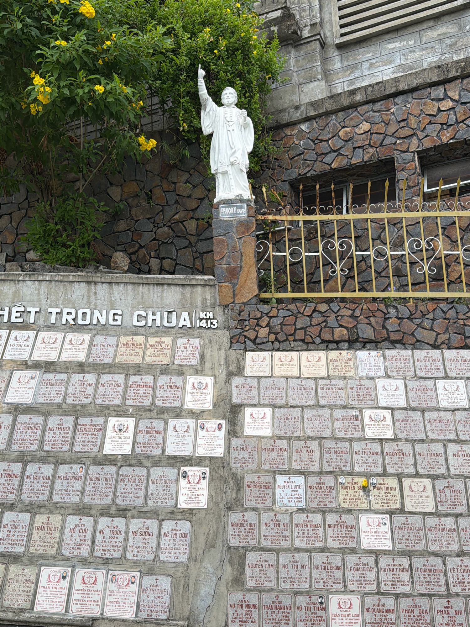 The image shows a monument in Nha Trang, Vietnam, dedicated to those who died for the country. The monument is a wall with many stone tablets inscribed with names and dates, likely representing individuals who perished in the Vietnam War. A white statue of a religious figure, possibly a saint, stands at the top of the monument, with its right arm raised and index finger pointing upwards. Behind the monument, there is a stone wall and a yellow wrought-iron fence. Yellow flowers bloom on a green bush to the left of the statue. The overall scene appears to be a memorial site.