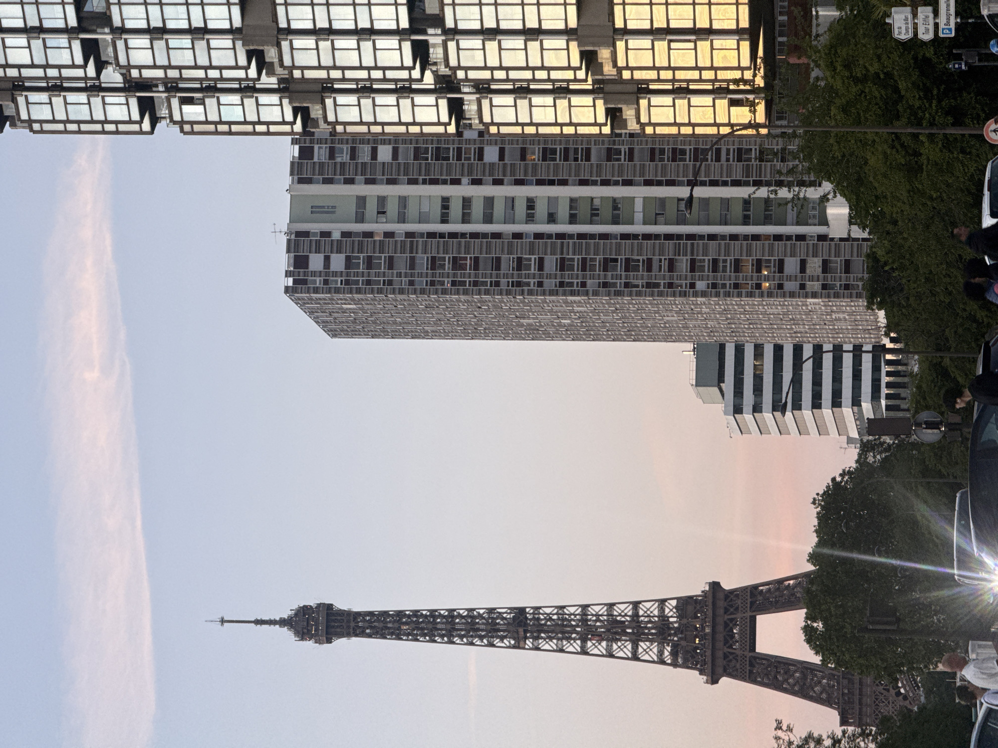 The image shows the Eiffel Tower in Paris, France, with modernist buildings in the background. The tower is silhouetted against a pale sky, likely at dawn or dusk. Above it is a tall building with a repetitive pattern of windows. Another building with a unique boxy architectural style with a reflection is present near the top of the frame. Cars and foliage can be seen at the side. People are standing around, and there are street signs visible as well.