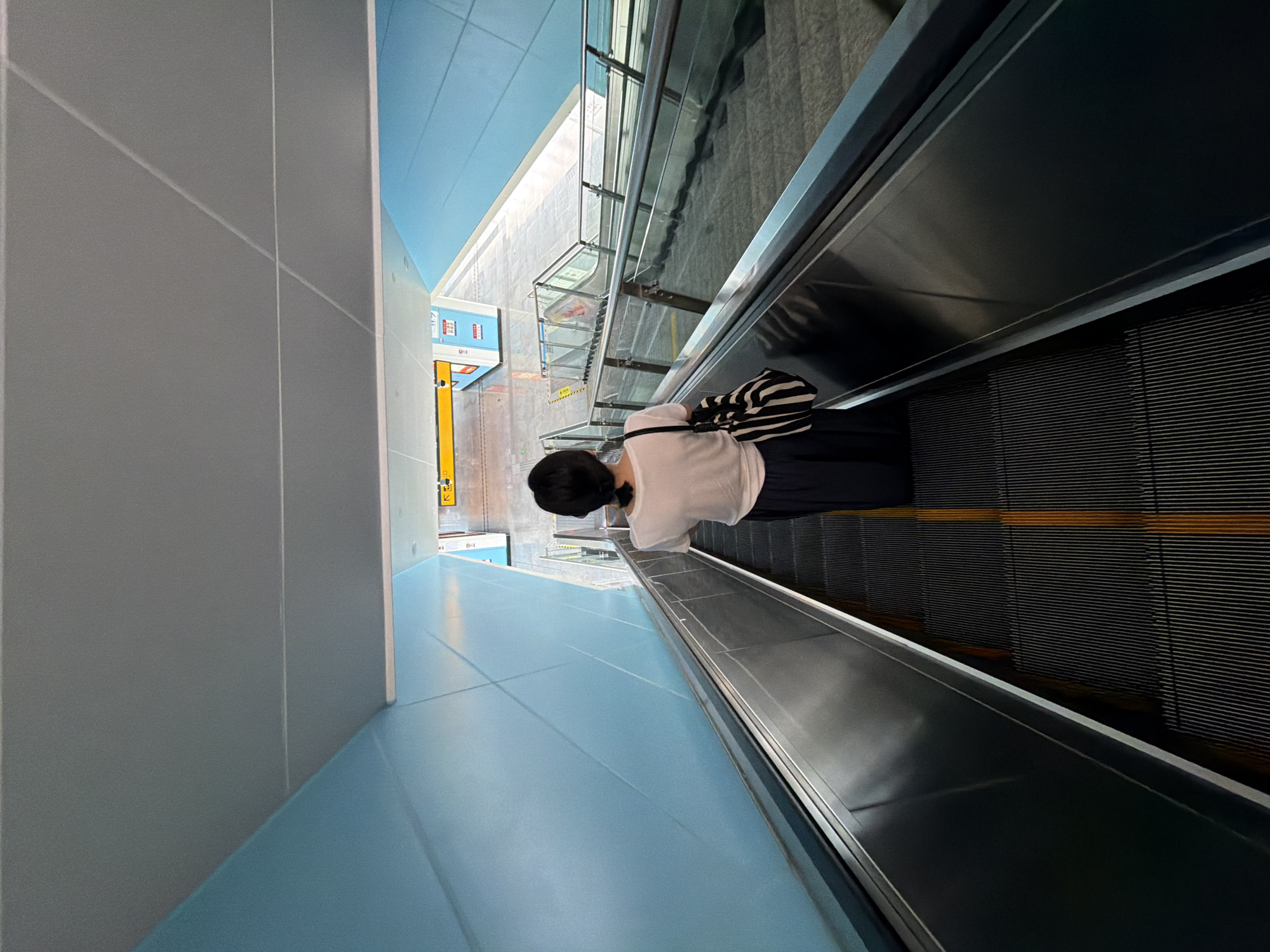 A person walks down a downward-moving escalator. The person is viewed from behind, wearing a white t-shirt and dark pants. They are carrying a black and white striped bag over their left shoulder. The escalator is metallic with black steps and yellow safety lines. The walls of the surrounding area are a light blue color. In the background, there is a street or platform with signs and other escalators. The scene appears to be in a public transportation hub, possibly a subway station or airport, in Jiangjin, China.