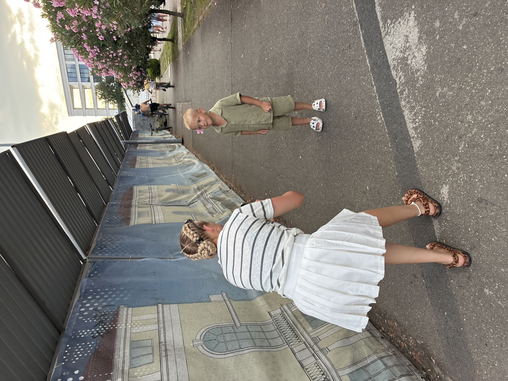 The image depicts a street scene in Sochi, Russia. A woman, viewed from behind, is crouching down and holding a smartphone, appearing to take a photograph of a young boy. The woman is wearing a striped t-shirt and a white pleated skirt, with her hair styled in braids. The boy, standing on the asphalt path, is wearing a khaki-colored short-sleeved shirt and matching shorts. He has a pink flower tucked behind his right ear. The background shows a long, blue mural depicting classical architecture, likely a temporary barrier. Beyond the mural, several people are cycling, and a modern white building with balconies is visible, surrounded by trees with pink blossoms. The sky above is cloudy with hints of sunset light. The ground is an asphalt path with a distinct black stripe running across it.