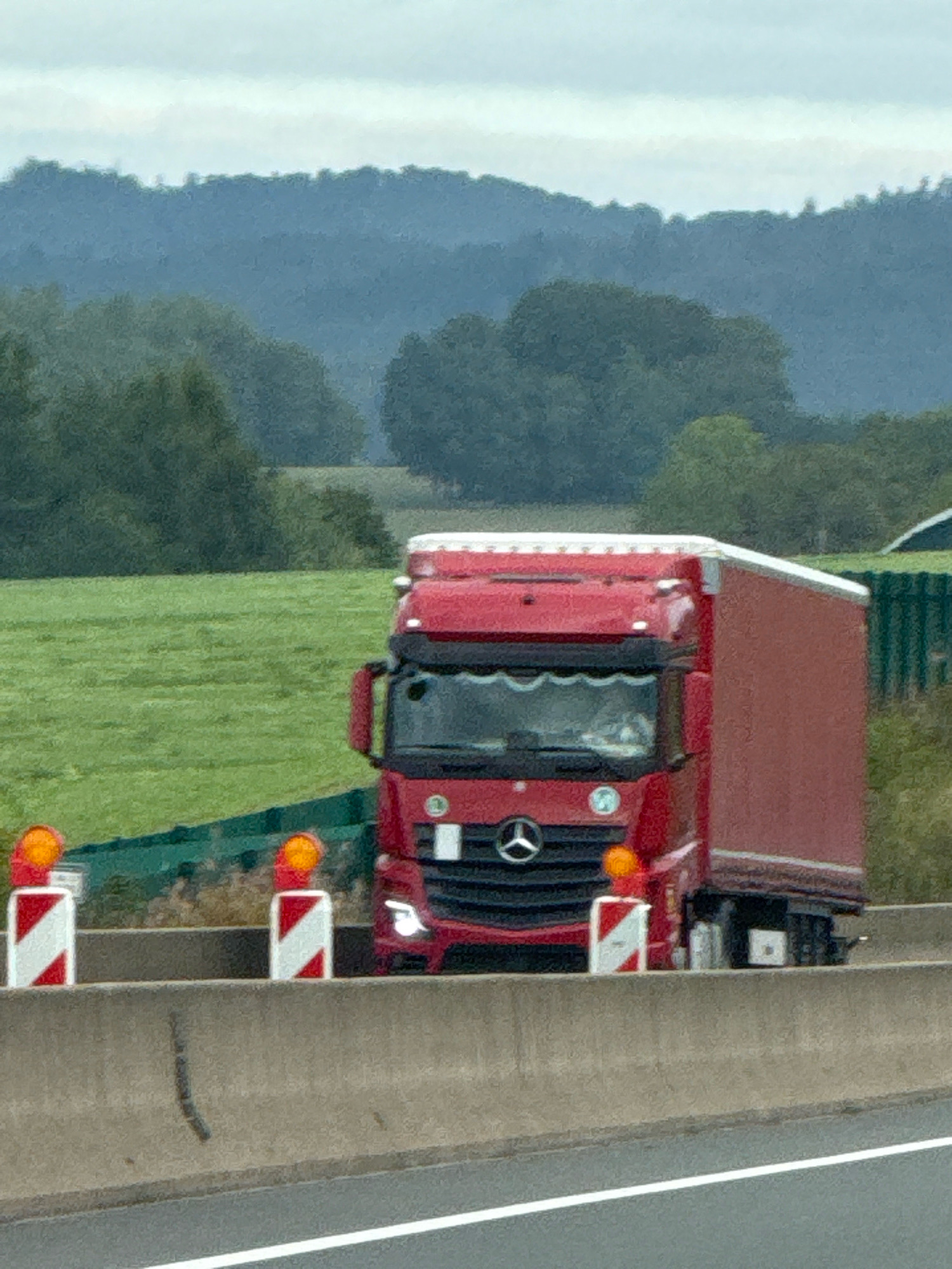 A red semi-truck is driving on a highway with a concrete barrier in the foreground and hills in the background. The truck has a trailer and appears to be in motion. There are several orange traffic cones with reflective white and red stripes on the barrier. The sky is overcast. The location is likely in or around Bad Lauterberg im Harz, Germany, as indicated by the presence of rolling hills typical of the Harz region.