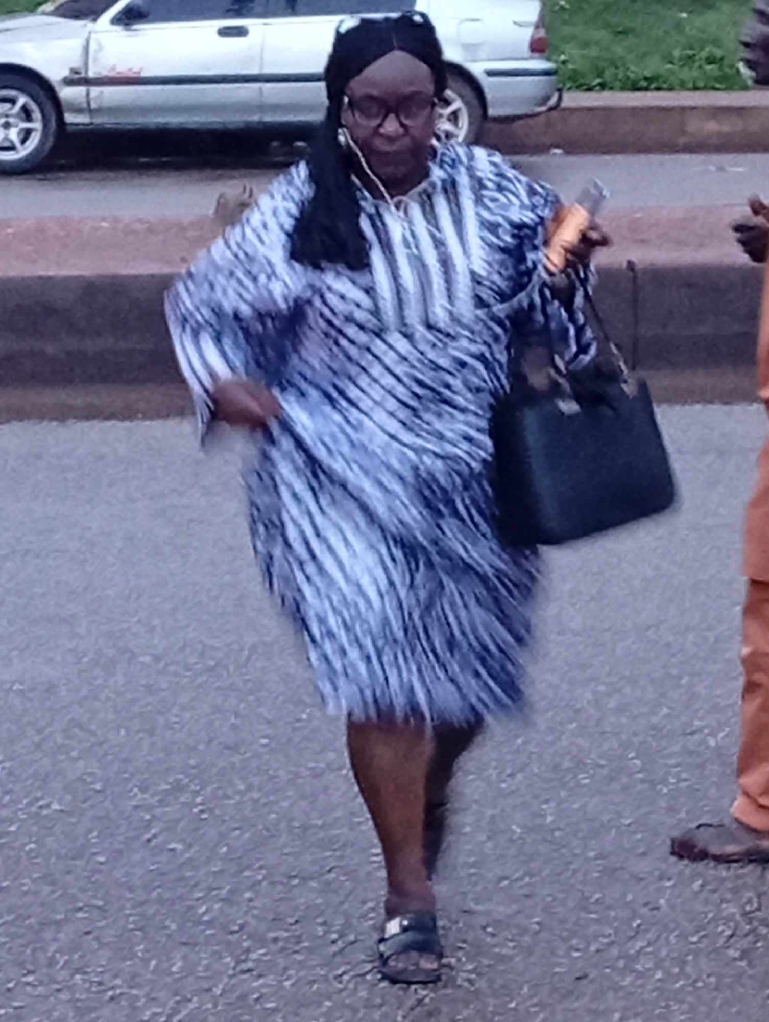 A woman is captured mid-stride on a paved surface, likely a road or sidewalk in Jos, Nigeria. She is wearing a dark headscarf, glasses, and a long, flowing dress with a striking blue and white striped pattern. She has a black handbag slung over her left shoulder and is holding a bottle or can in her right hand, which appears to be an orange drink. Her feet are clad in dark sandals. In the background, a silver car is parked, and a patch of green vegetation is visible. The scene suggests a casual urban environment, possibly near a street.