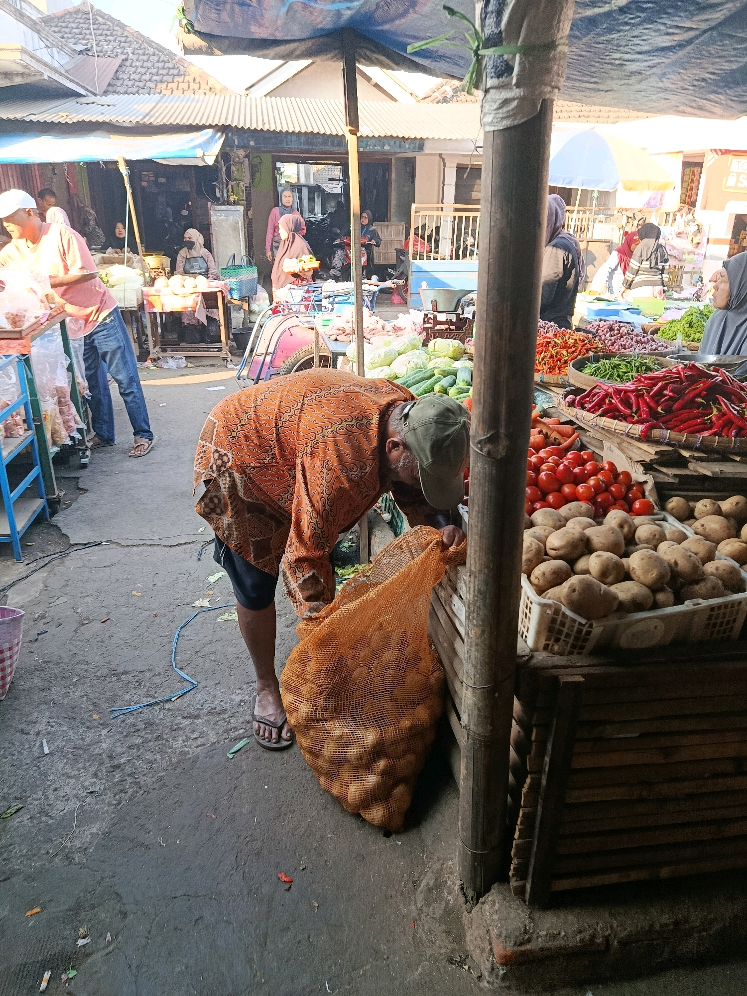 An outdoor market scene in Gondanglegi, Indonesia, shows a vendor, an elderly man wearing a patterned orange batik shirt, a green cap, and dark shorts, bent over and reaching into a large mesh bag of potatoes. He is standing on a paved ground, with a blue rope trailing nearby. To his right, a wooden stall displays various fresh produce, including a basket overflowing with red chili peppers, a crate of tomatoes, and a bin of potatoes. In the background, other market-goers are visible, some behind stalls and others walking through the market. The market appears to be covered by tarps, suggesting it is a bustling, possibly open-air marketplace typical of Indonesian towns.