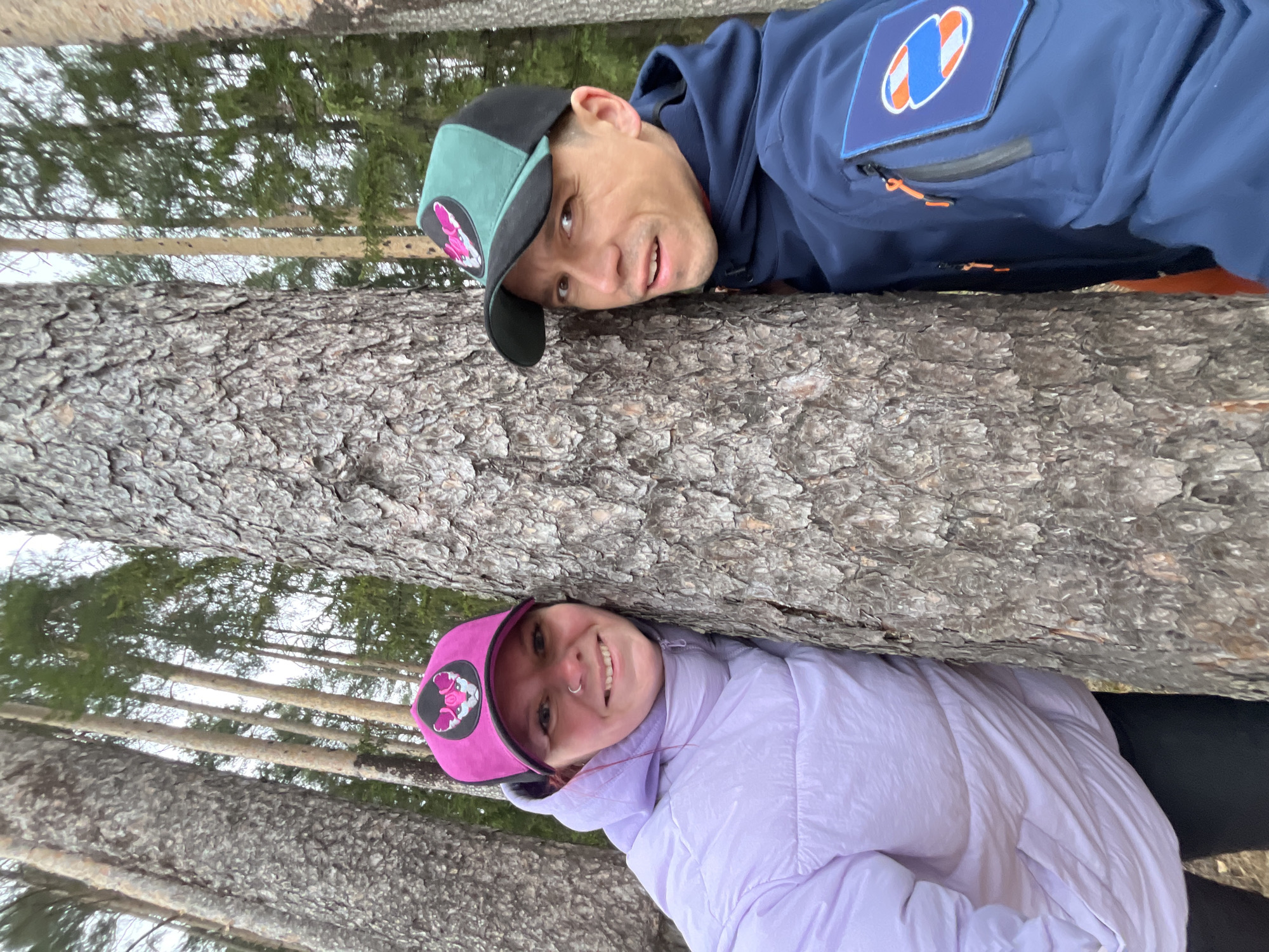 Two people, a man and a woman, are positioned between what appears to be two large trees. The man is above, wearing a dark blue jacket with an orange accent and a patch, and a two-tone black and green cap with a pink logo. The woman is below, wearing a light purple jacket and a pink cap with a black logo. Their faces are visible, with both looking at the camera. The background shows an out-of-focus forest scene with trees. The location is in a forest, where it is possible to take a picture of such things. The city of Chaikovsky, Russia is not indicated.