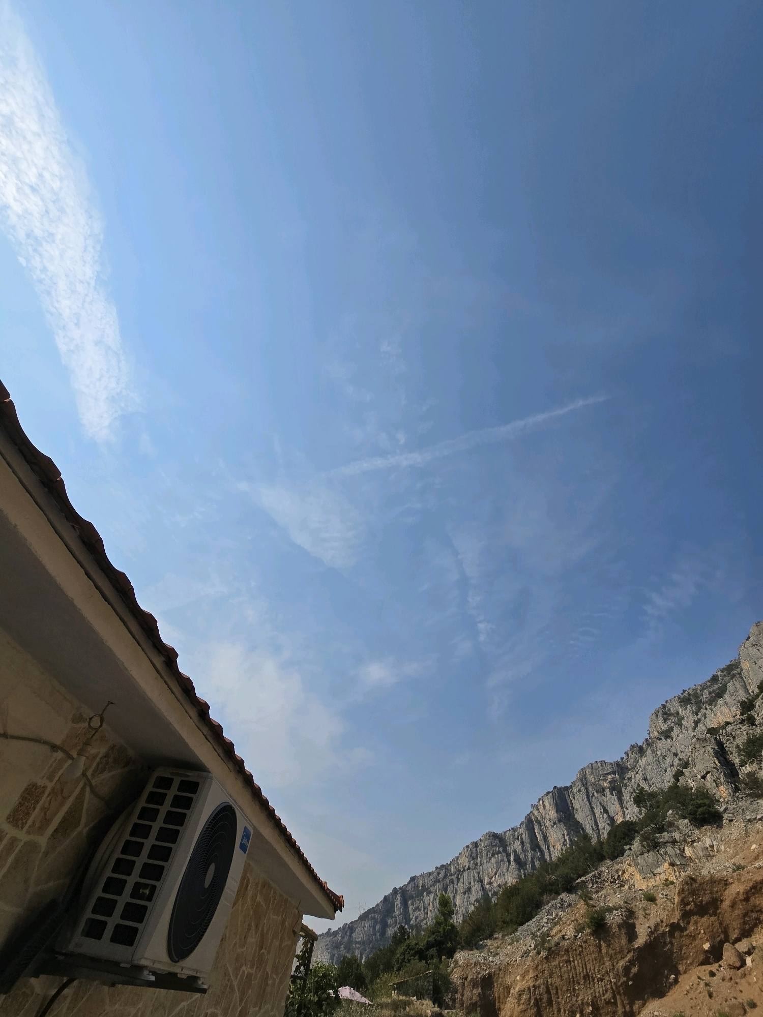 An outdoor, low-angle shot captures a corner of a building in the foreground on the left, with an outdoor air conditioning unit mounted on its stone-tiled wall. The building's red-tiled roof is visible. The background features a steep, rocky mountainside with sparse vegetation, under a bright blue sky with scattered wispy clouds. The scene is indicative of a mountainous or hilly region, likely on the outskirts of a city such as Podgorica, Montenegro, known for its proximity to such natural landscapes. No people are present in the image.