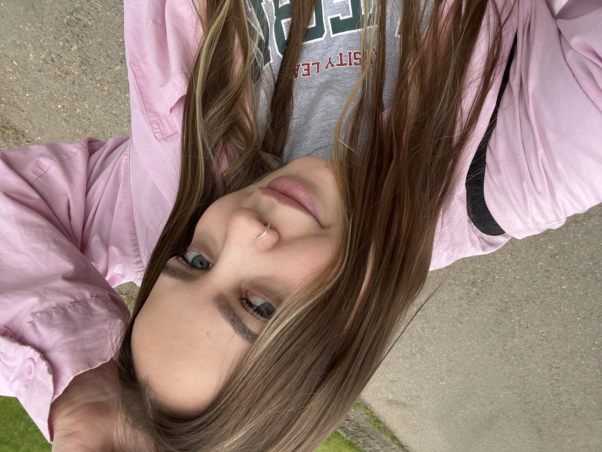 A young woman with light brown hair and green eyes is centered in an upside-down close-up shot. She is wearing a pink button-down jacket over a gray t-shirt with green and red lettering. A silver nose ring is visible. The background consists of a concrete surface and patches of green grass. The location, Novopokrovskaya, Russia, is not discernible from the visual content.