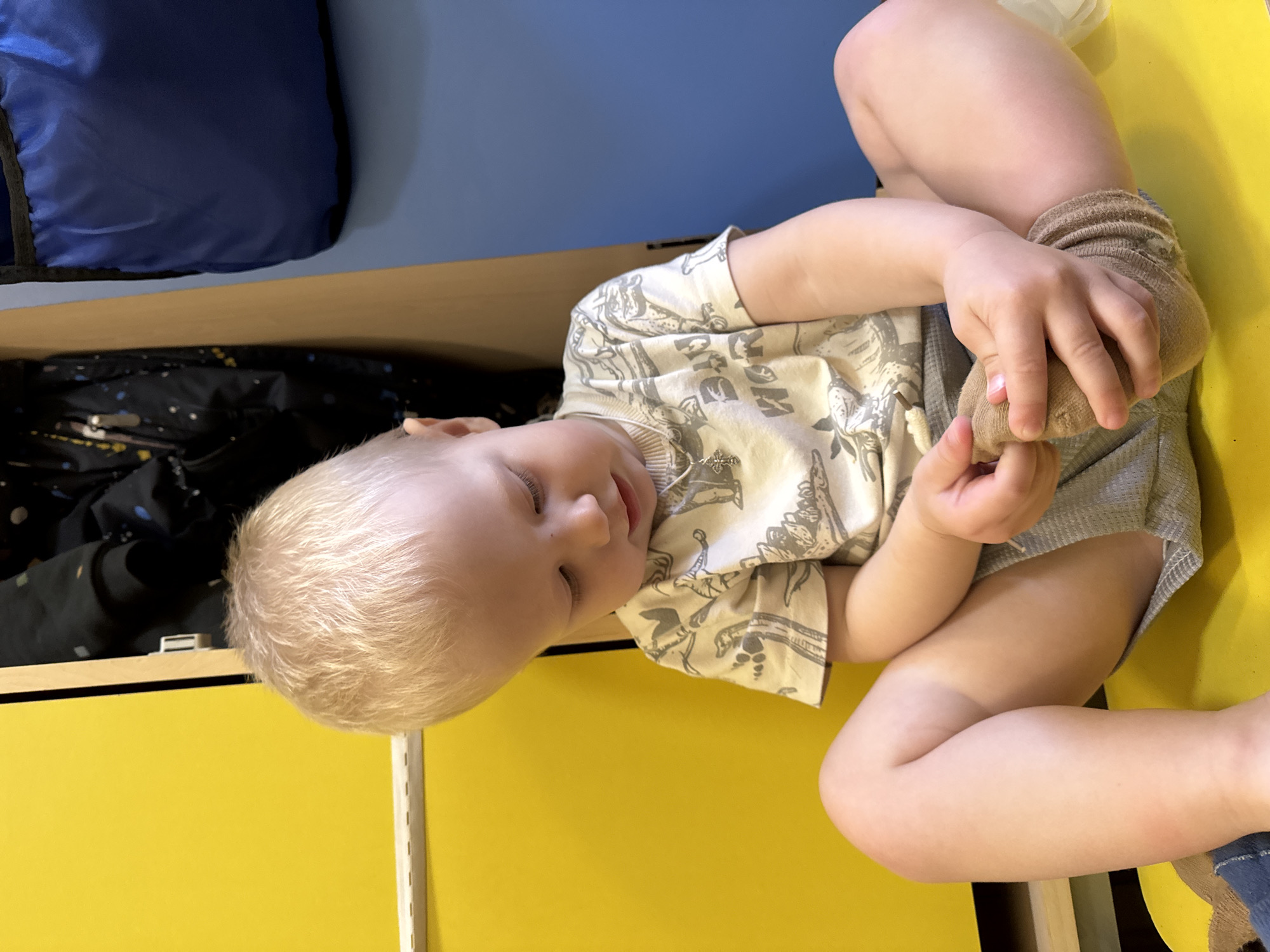 A young child, likely a toddler, is depicted lying on their back, with their head positioned towards the left side of the frame. The child has very light blonde hair and appears focused on their own foot, which they are holding with both hands. One foot wears a light brown, slightly bunched sock. The child's eyes are either closed or cast downwards intently, and their mouth is slightly ajar, suggesting a moment of quiet concentration or play.
They are wearing a light-colored (possibly off-white or cream) short-sleeved top adorned with grey patterns that resemble dinosaurs or other stylized figures, and grey shorts.
The child is resting on a bright yellow surface, which appears to be the top or a large panel of a piece of furniture, possibly a bed or a low cabinet. To the child's right (our upper left, given the original image's rotation), there is an open compartment revealing a dark fabric item, possibly a bag or folded clothing, with faint light-colored patterns. Above this compartment is a light brown wooden edge. The wall behind the furniture is a solid, medium blue color. A dark blue fabric object, resembling a bag or cushion, is visible against the blue wall in the upper left corner. To the child's left, a vertical white metal rail, possibly part of a shelving or bed frame system, runs along the edge of the yellow surface.
The scene appears to be indoors, under ambient lighting, creating a soft shadow. The bright yellow furniture and blue wall contribute to a cheerful, child-friendly environment. There is no visible text on the child's clothing or in the immediate surroundings that is clearly legible. The overall impression is one of a calm, everyday moment of a child engaged in self-play. The location context is Chaikovsky, Russia.