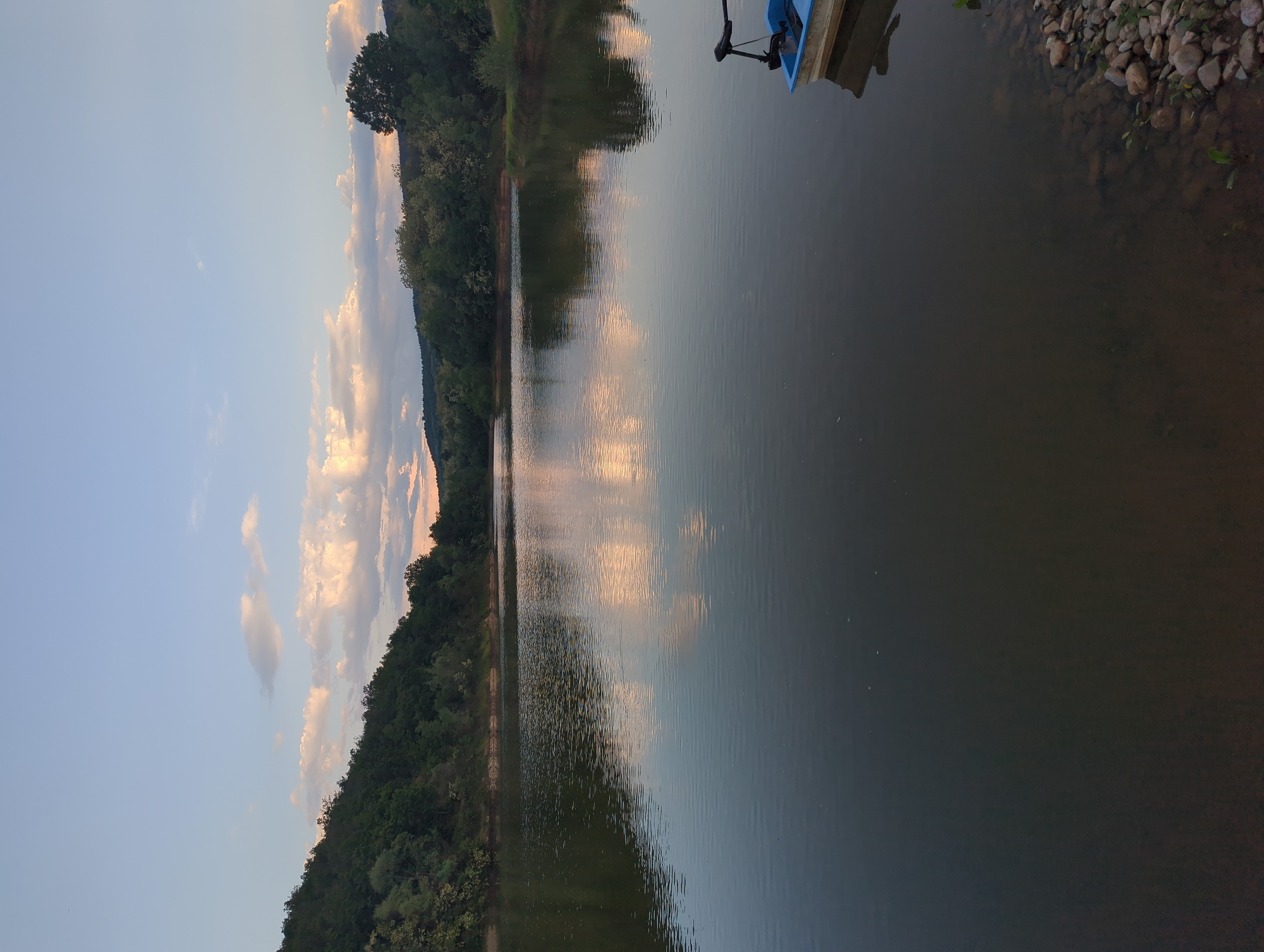 The image shows a landscape, likely at a lake or river. On the right, there's a shoreline with rocks and some vegetation. A portion of a blue boat or dock is visible in the top right corner. The water reflects the sky and possibly trees on the opposite shore. The left side of the image reveals a forested hillside, and the sky is pale blue with some clouds that have a pink hue, indicating it may be near sunrise or sunset.
I lack the location information so cannot provide more specifics or confirm if it is Slavovitsa, Bulgaria.