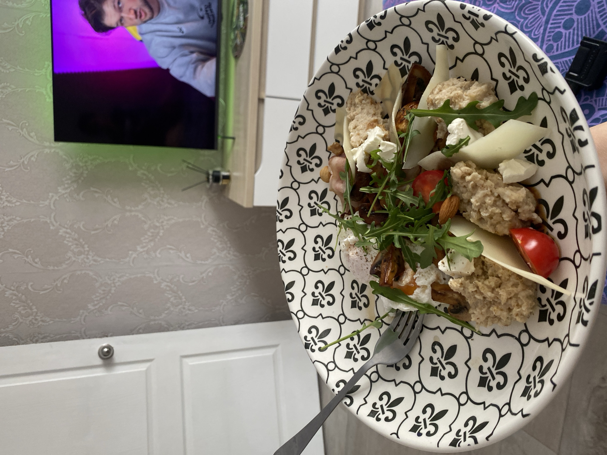 The image shows a plate of food held in front of a TV screen, likely indoors. The plate features a black and white patterned design and contains what appears to be a salad with ingredients such as grains, cheese, tomatoes, almonds, arugula, and perhaps some kind of cooked egg. A fork is positioned on the plate, suggesting an intention to eat. The TV screen displays a man in a grey sweatshirt. The backdrop includes a wall with patterned wallpaper and white cabinets. There is no definitive information in the image to confirm a location within the city of Chaikovsky, Russia.