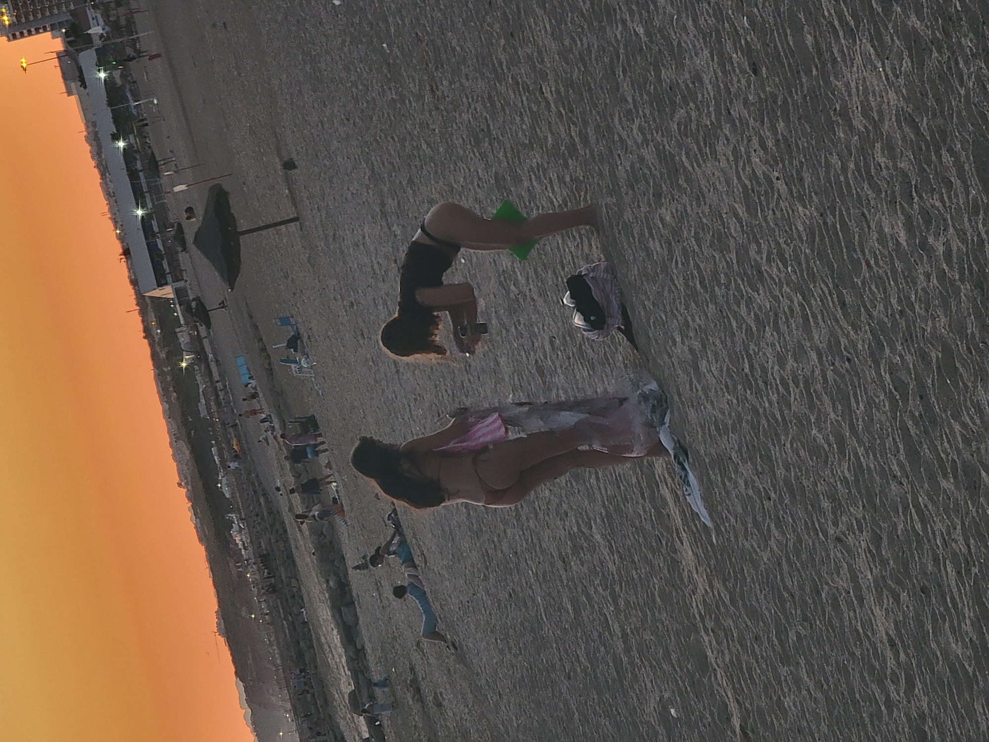 The image depicts a beach scene in Quarteira, Portugal, during sunset. The sky is illuminated with an orange hue, characteristic of the twilight hours. In the foreground, two women are present on the sandy beach. The woman on the left is wearing a bikini and is holding a rolled-up towel or sheet. To her right, another woman in a bikini is bending down, holding a smartphone and a green object, possibly a bag or a towel. Several beach umbrellas and lounge chairs are scattered across the beach, and in the background, there are more people enjoying the beach and distant buildings. The overall atmosphere suggests a relaxed evening by the sea.