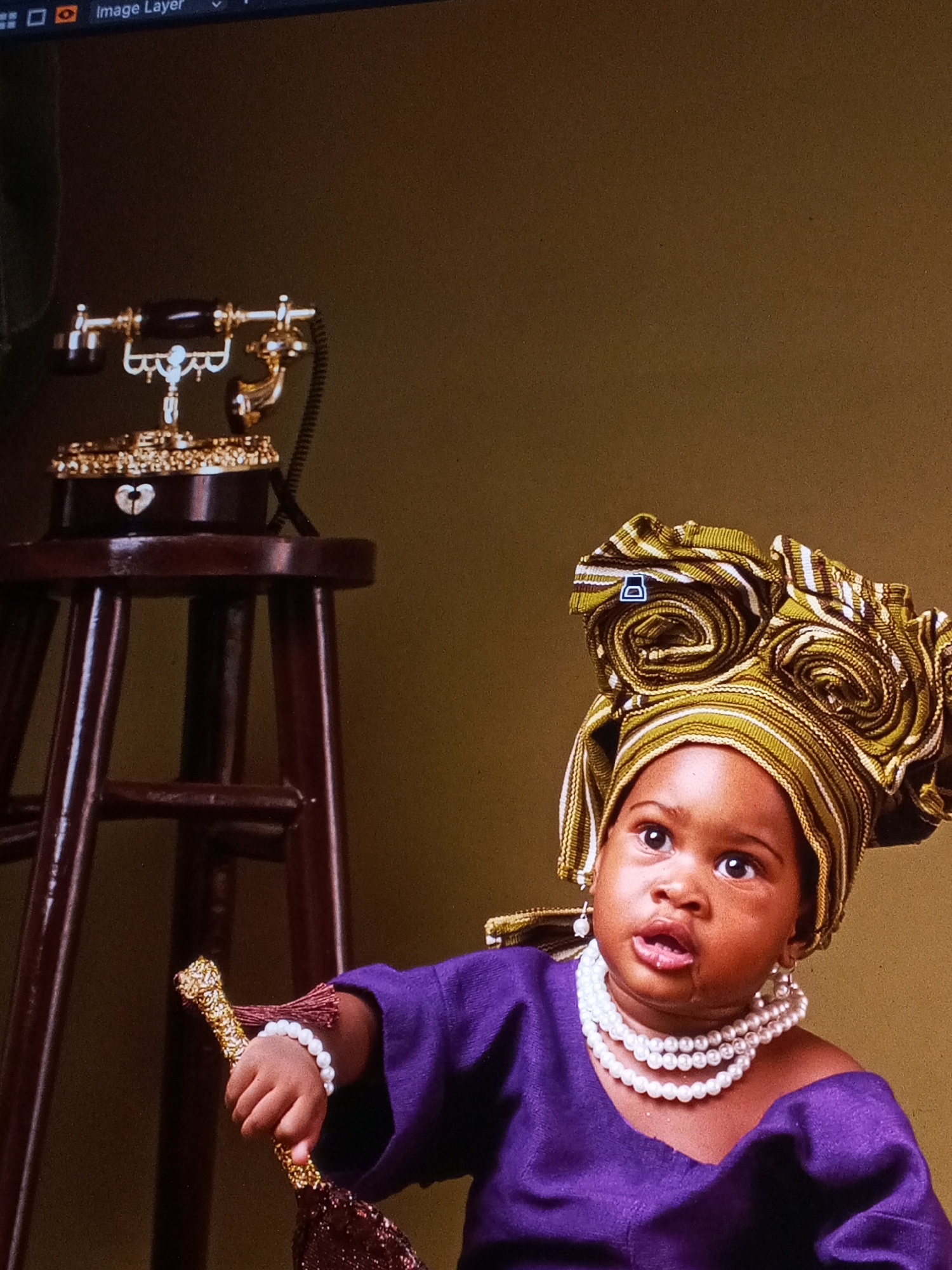 A baby in traditional Nigerian attire is the primary subject. The baby wears a green and yellow striped head wrap styled into a rosette shape, multiple strands of white pearls around the neck, a matching pearl bracelet, a purple garment, and small dangle earrings. The baby is holding a gold ornate whisk. To the left of the baby is a dark wood stool with an antique gold and brown rotary phone sitting on it. The background is a solid olive-green color. There is no other information available to confirm if it's taken in the city of Badagry, Nigeria.