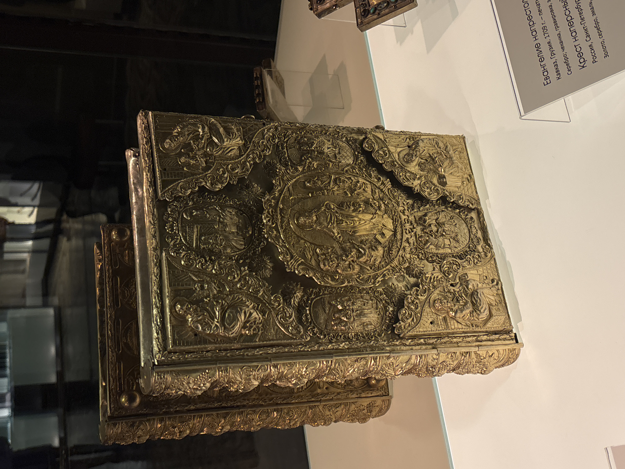 The image displays a detailed, ornate gold-covered gospel book resting on a lower shelf, likely a display case. The book cover features intricate carvings and embossed imagery. A descriptive plaque is visible in the background, likely providing information about the book, including its origin (Caucasus, Georgia, 1709) and materials (silver). The text mentions it as an "Evangelie naprestol" (Gospel upon the altar) which suggests it is from a religious context. In the same placard it mentions "Krest napresny" (Cross worn over chest) from Russia, St. Peterburg, which indicates they are exhibiting religious artifacts in a museum or a historical display setting, potentially in Moscow, Russia, as the text is in Russian. The environment appears dimly lit, indicative of a museum or similar controlled setting designed to protect delicate artifacts.
