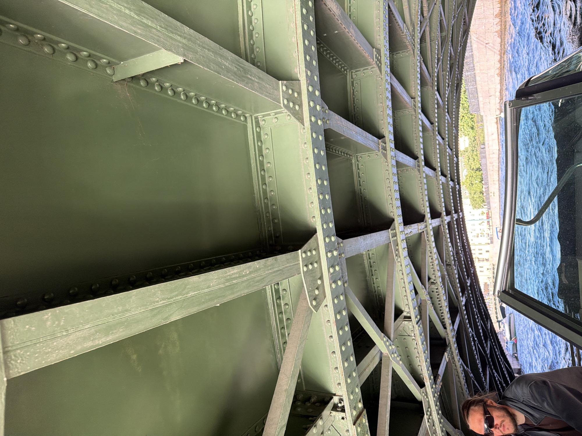 The image shows the underside of a green metal bridge with multiple levels of beams and rivets. Below the bridge, a body of water is visible, with a boat in the foreground. A man wearing sunglasses and a dark jacket is seated in the boat, looking to the left. In the background, beyond the bridge, buildings and trees are visible along the waterfront. The overall scene suggests a view from a boat passing under a bridge in an urban setting.