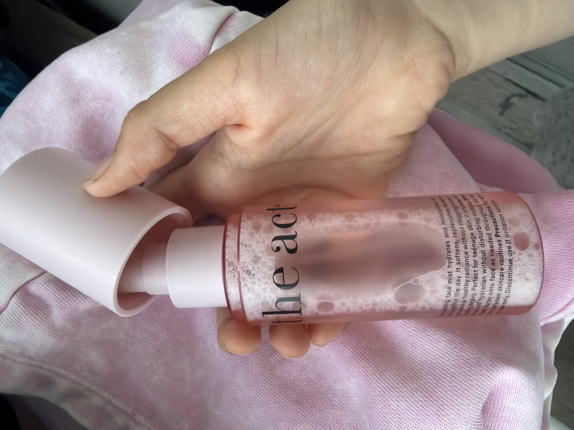 A hand, partially visible, holds a clear pink plastic bottle containing a foamy liquid. The bottle has a white cap, which is partially removed. Black text is printed on the bottle, the visible portion reading "the act" and some smaller text below. The bottle is resting against a pink fabric item, likely clothing. The background is out of focus, showing some darker colors and a hint of a textured surface. The image is taken from a close-up perspective.