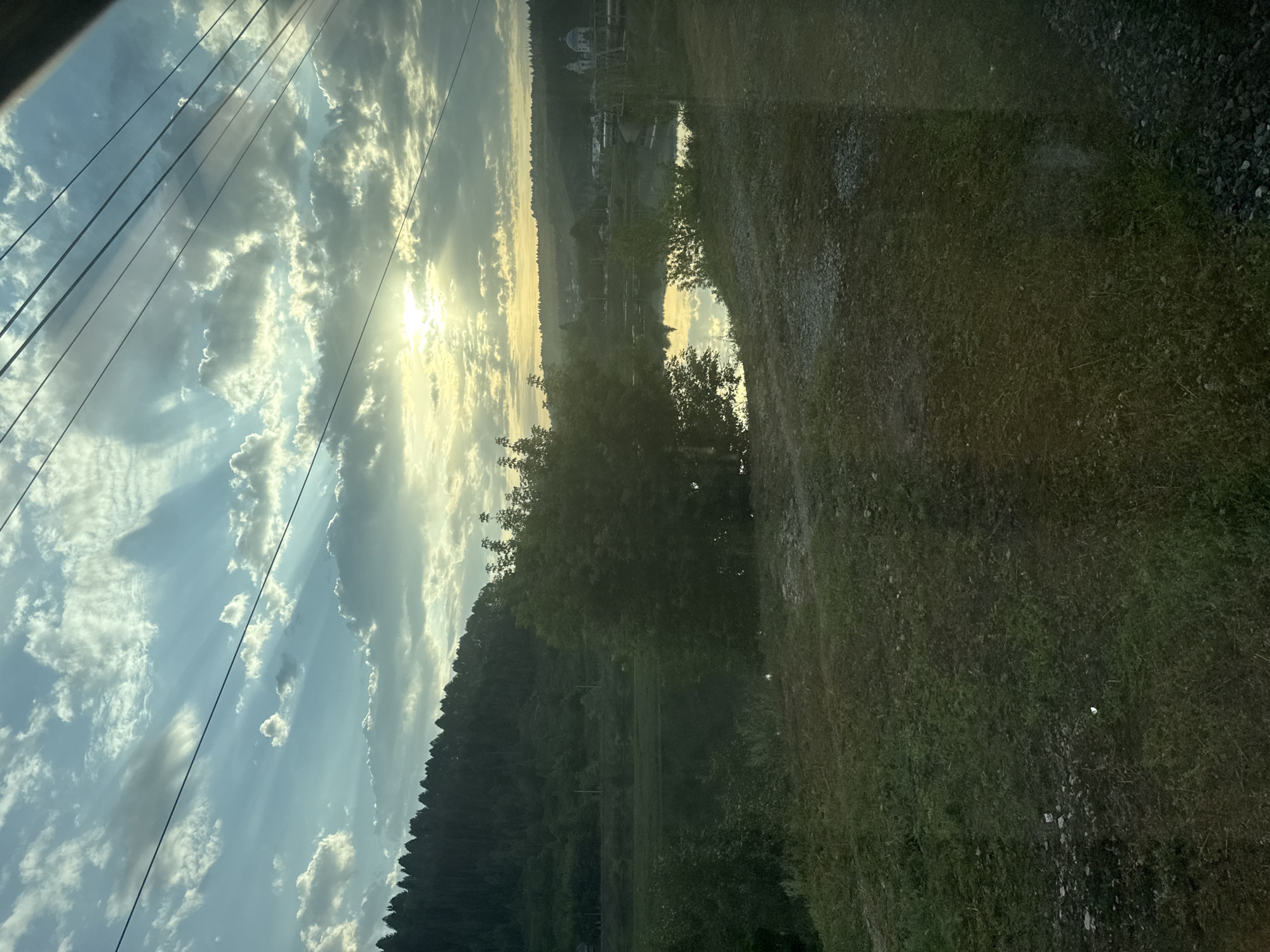 This image depicts a serene landscape scene during what appears to be sunrise or sunset, with the sun's rays piercing through a cloudy sky. The foreground is dominated by grassy terrain with scattered gravel and some patches of darker soil. A body of water, likely a river or lake, is visible in the middle ground, reflecting the sky and surrounding trees. On the far bank of the water, a dense forest covers a hillside. In the distance, across the water and beyond the trees, a settlement can be seen with buildings, including a prominent white structure with domes, possibly a church or monastery. Overhead, power lines crisscross the frame, suggesting the presence of infrastructure near the natural landscape. The overall atmosphere is tranquil and bucolic. The location is identified as Minyar, Russia.