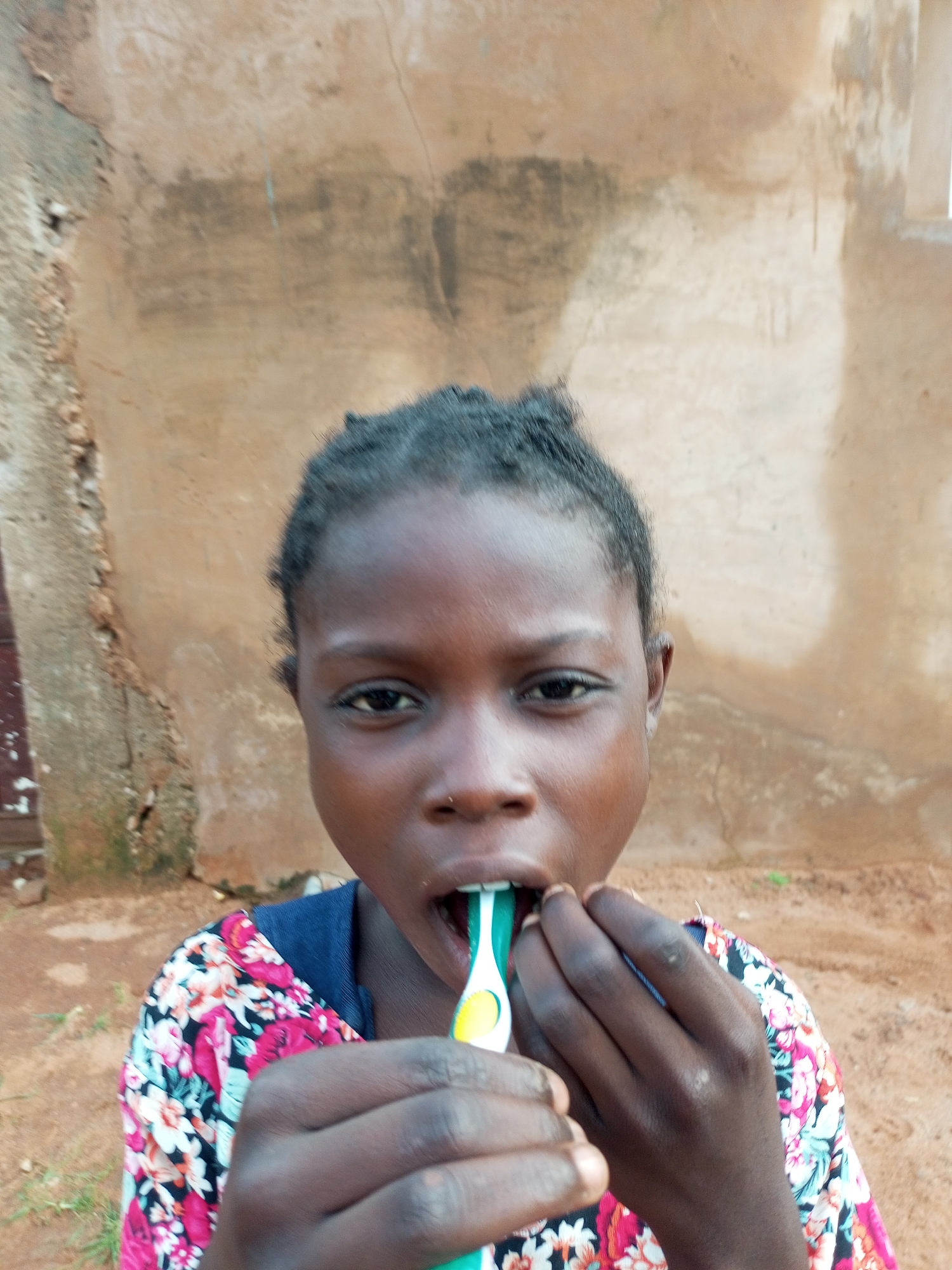 A young girl with dark skin and braided hair is shown brushing her teeth outdoors. She is holding a toothbrush with green and yellow bristles in her right hand, and her left hand is near her mouth. She is wearing a colorful shirt with a floral pattern and a blue collar. The background appears to be a rough, sandy ground with a textured, light brown wall behind her. The wall has some dark stains and appears to be made of mud or stucco. A doorway or window opening is visible on the right side of the wall. The lighting suggests it is daytime. The overall impression is of a candid moment of personal hygiene in a rustic environment. The location context is Tempure, Nigeria. There is no visible text in the image.