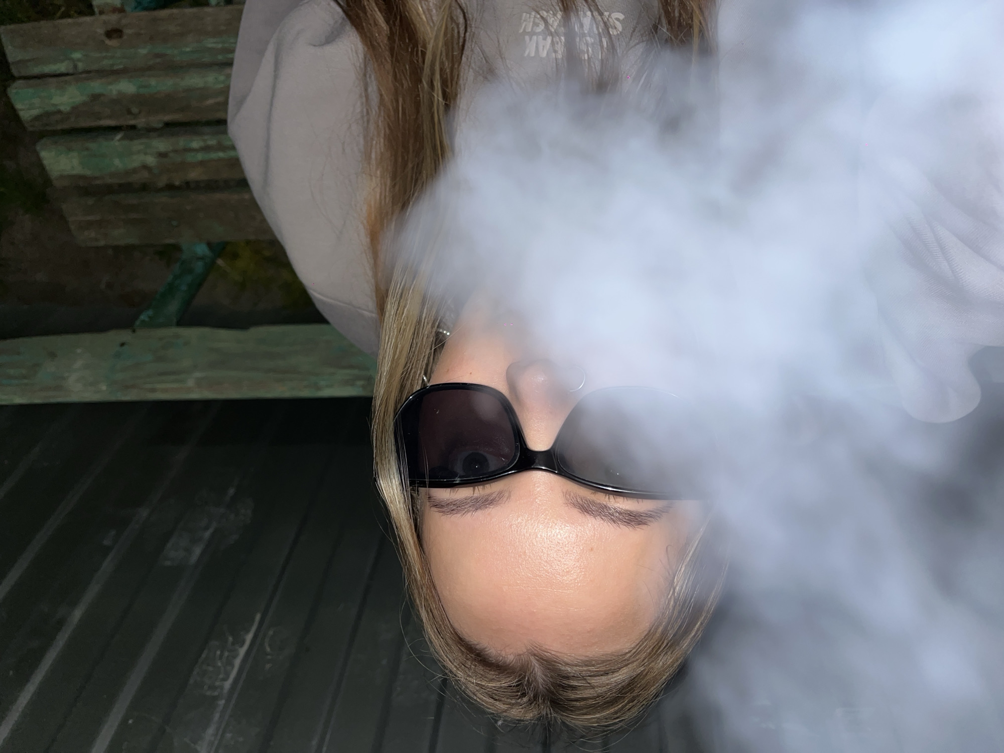 A person with blonde hair and sunglasses is partially obscured by smoke. The person is wearing a light-colored hooded sweatshirt. The background includes a wooden bench. The image appears to be taken in Novopokrovskaya, Russia.