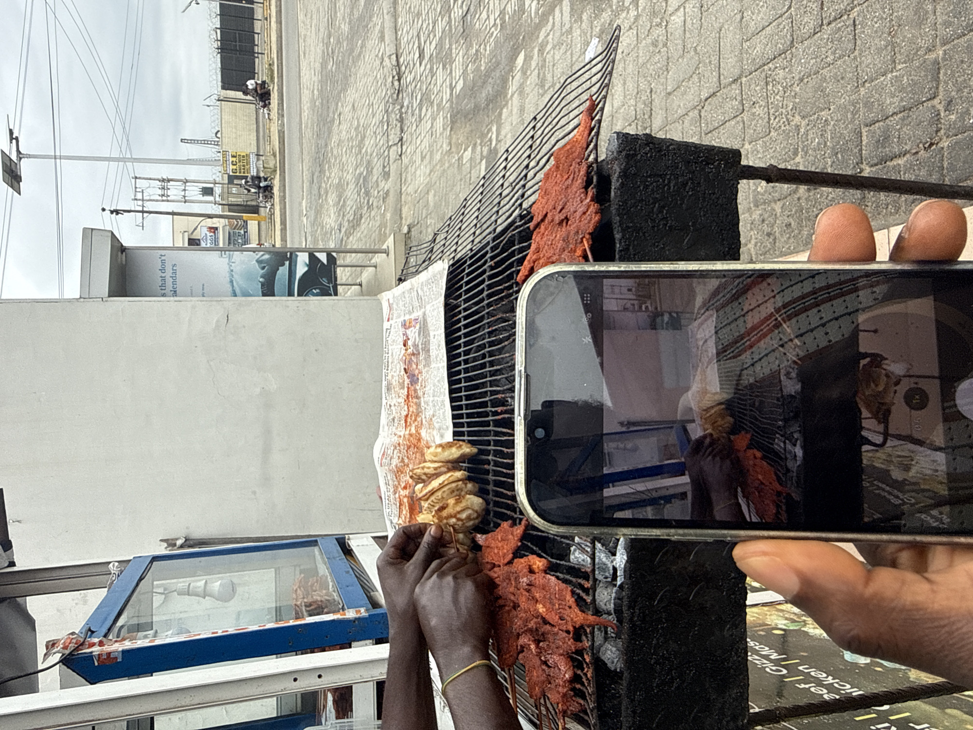 In Ado, Nigeria, a close-up shot focuses on a street food vendor preparing meat skewers over a charcoal grill. The foreground shows a hand holding a smartphone, capturing the scene. The grill, made of dark metal, holds rows of red, marinated meat on skewers and some grilled plantains. In the background, a blue-fronted display case, likely for food storage, is visible, along with a white building. Beyond that, a street scene unfolds with pedestrians, motorcycles, and overhead electrical wires, under a cloudy sky. A sign advertising "G.C.E. REVISION MASTER" is visible on a yellow board in the middle ground. The overall impression is of an outdoor food stall operating in an urban environment.