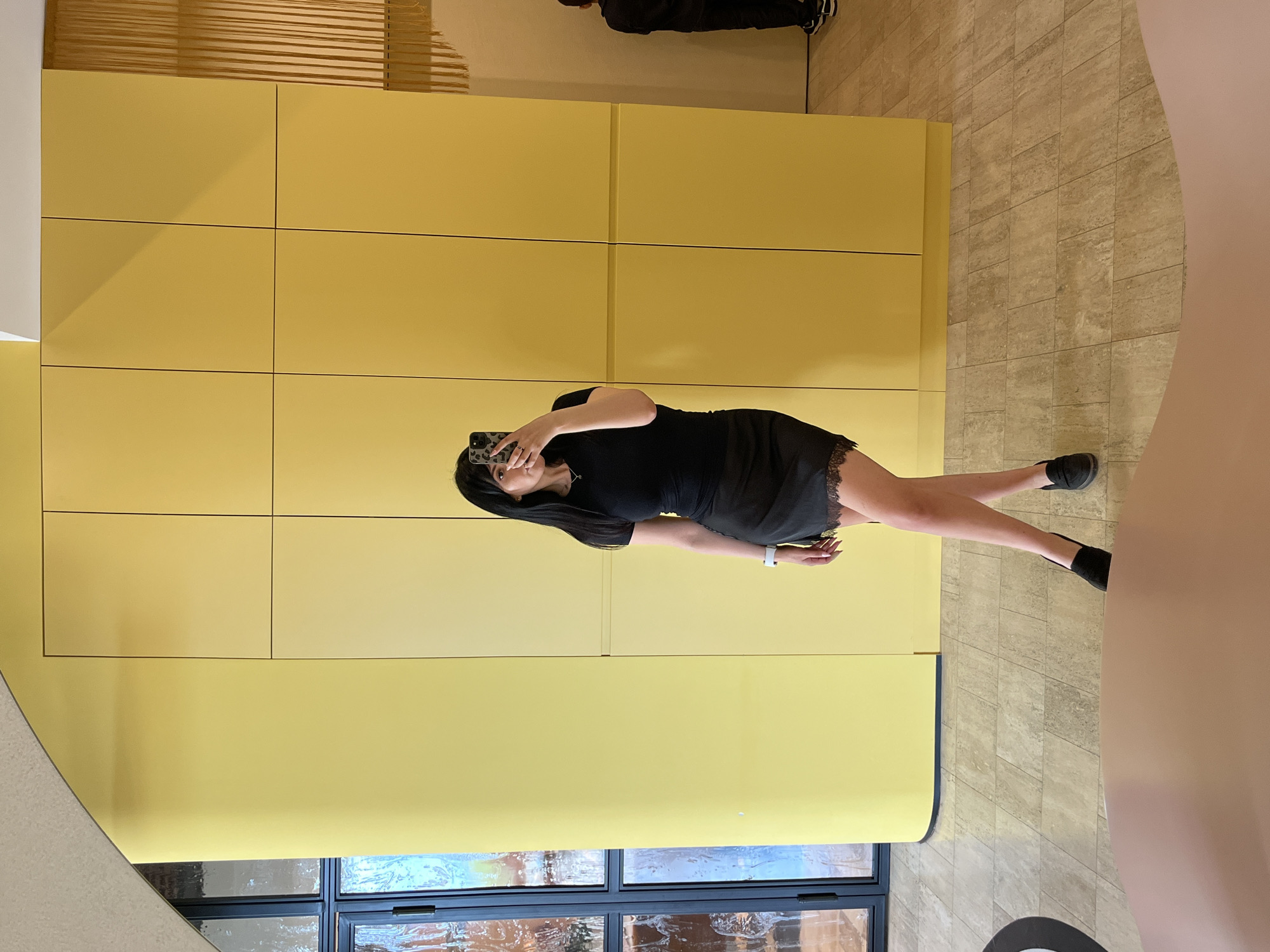 A woman is standing in front of a yellow wall, taking a picture of herself with a cell phone. She is wearing a black dress and black shoes. The wall is made of rectangular panels. There are windows at the bottom of the wall. The location is likely in an area with modern architecture, and judging from the presence of Russian text in other related images, the city of Sirius, Russia, is possible. The floor is made of light-colored tiles.