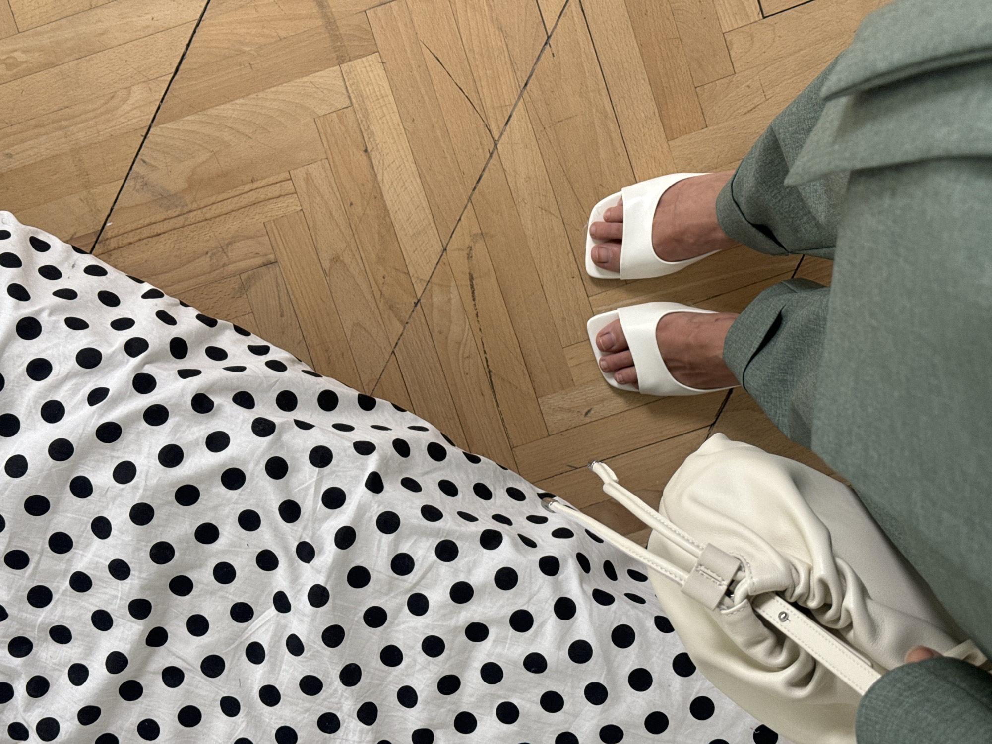 A person's legs and feet, wearing white slide sandals and green pants, are shown from a top-down perspective. The person is standing on a wooden floor. To the left of the person is a white bag with a ruched design and a strap. Partially visible is a white fabric with black polka dots draped over the floor. There is no indication of the city of Krasnoyarsk, Russia, or any other specific location.